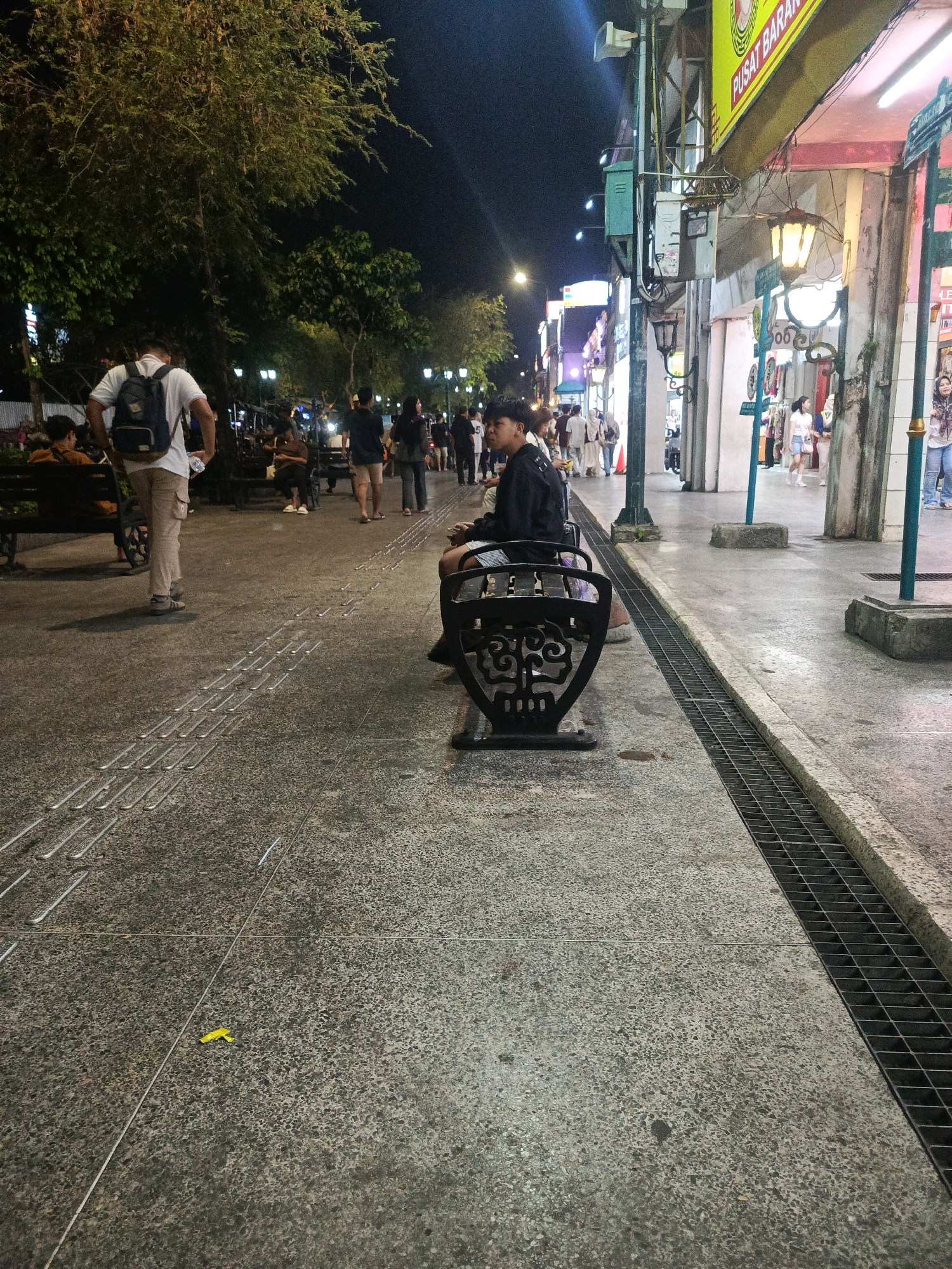 The image captures a nighttime scene on a pedestrian walkway in Yogyakarta, Indonesia, likely in a commercial area like Malioboro Street. The ground is paved with textured concrete, featuring a tactile paving system for the visually impaired. A metal grate runs along the edge of the walkway, bordering a drainage channel.
In the foreground, a person is seated on an ornate black bench, facing away from the camera. Further down the walkway, a person wearing a white shirt and a backpack is walking away, holding a water bottle. Several other individuals are visible, walking or sitting on benches, in the middle ground and background. Shops with illuminated signs and facades line the right side of the walkway, indicating a commercial setting. Trees with dense foliage are present on the left, providing shade and an urban green element. The overall atmosphere is that of a lively evening in a public space.