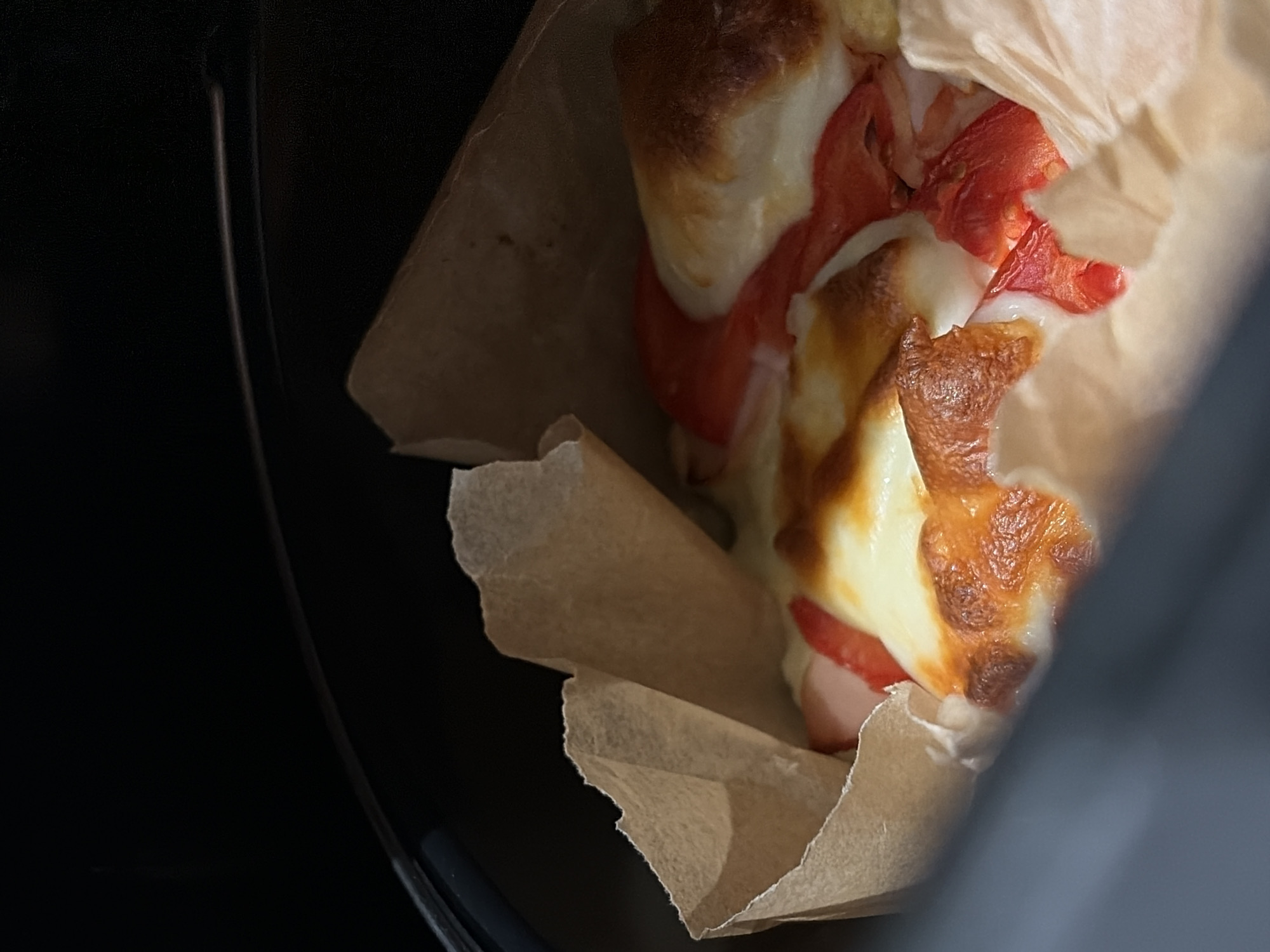 A close-up, high-angle shot reveals a savory baked food item partially wrapped in crumpled brown parchment paper. The food appears to be a small sandwich or pastry, generously topped with melted cheese that has developed a golden-brown, slightly charred crust in areas, indicating it was recently baked or toasted. Beneath the melted cheese, vibrant red slices of an ingredient, possibly tomato or a type of meat/pepper, are visible, adding a pop of color. The bread or base of the item is light in color, likely a soft roll or bun.
The food is positioned towards the right side of the frame, with the paper wrapping creating folds and texture around it. The background on the left and bottom is dark and indistinct, suggesting a shadowed interior space, possibly inside a vehicle or a dimly lit room. The lighting focuses on the food itself, highlighting the glossy texture of the melted cheese and the subtle colors of the ingredients. There is no visible text on the food, packaging, or in the background. The image captures a moment of consumption or preparation of a warm, comforting snack.