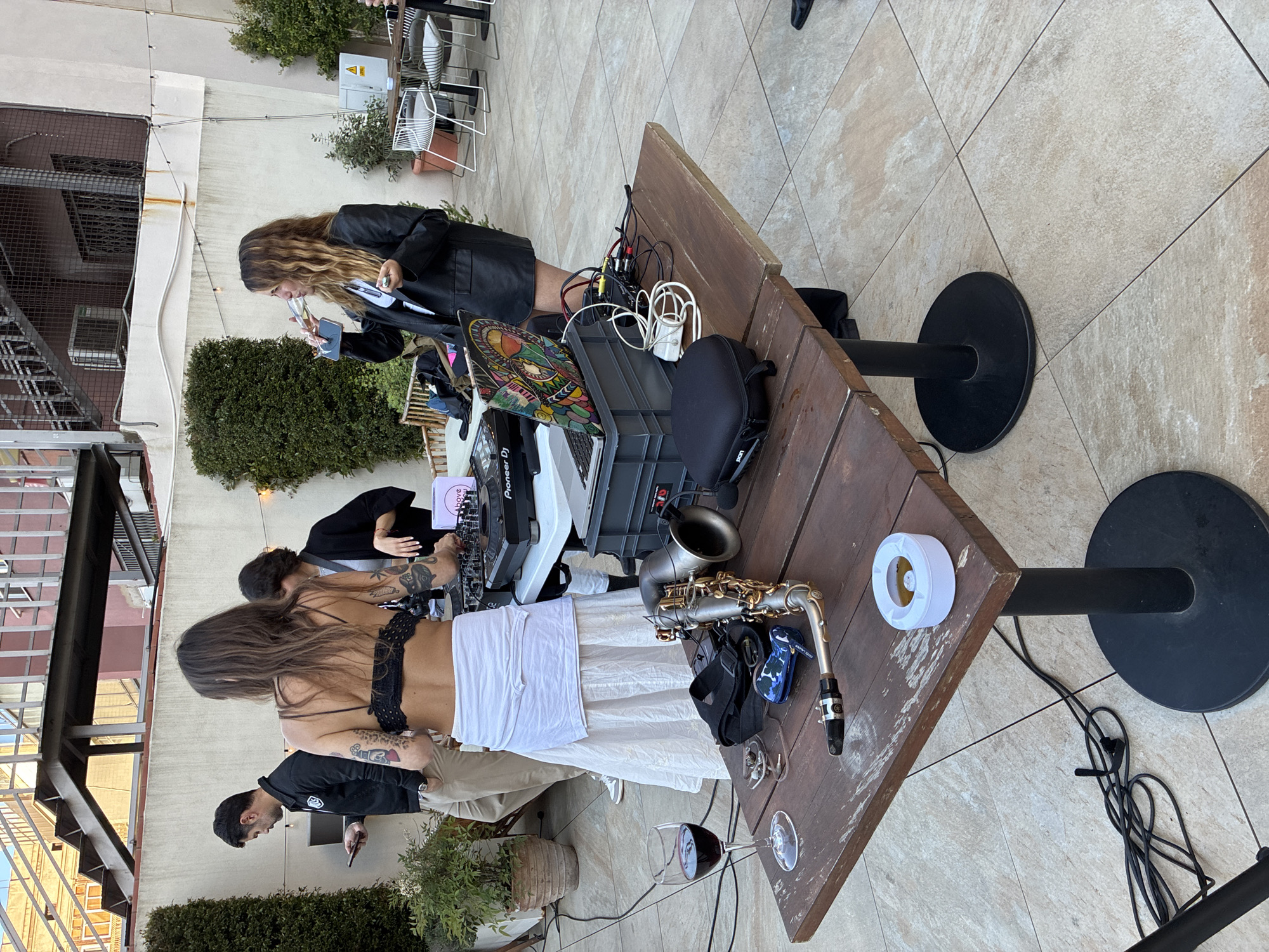 The image shows an outdoor performance setup on a patio in what appears to be Barcelona, Spain. A wooden table acts as a stage, equipped with a DJ setup including a Pioneer DJ controller, laptop, speakers, and assorted cables. A saxophone rests on the table next to a roll of white tape. A woman in a black leather jacket is near the DJ equipment. Another woman stands with her back to the camera, wearing a black top and white skirt. A man in a black shirt is also present. A glass of red wine sits near the edge of the table, along with a potted plant. A staircase and buildings are visible in the background, typical of urban architecture in Barcelona. The patio is paved with square tiles.