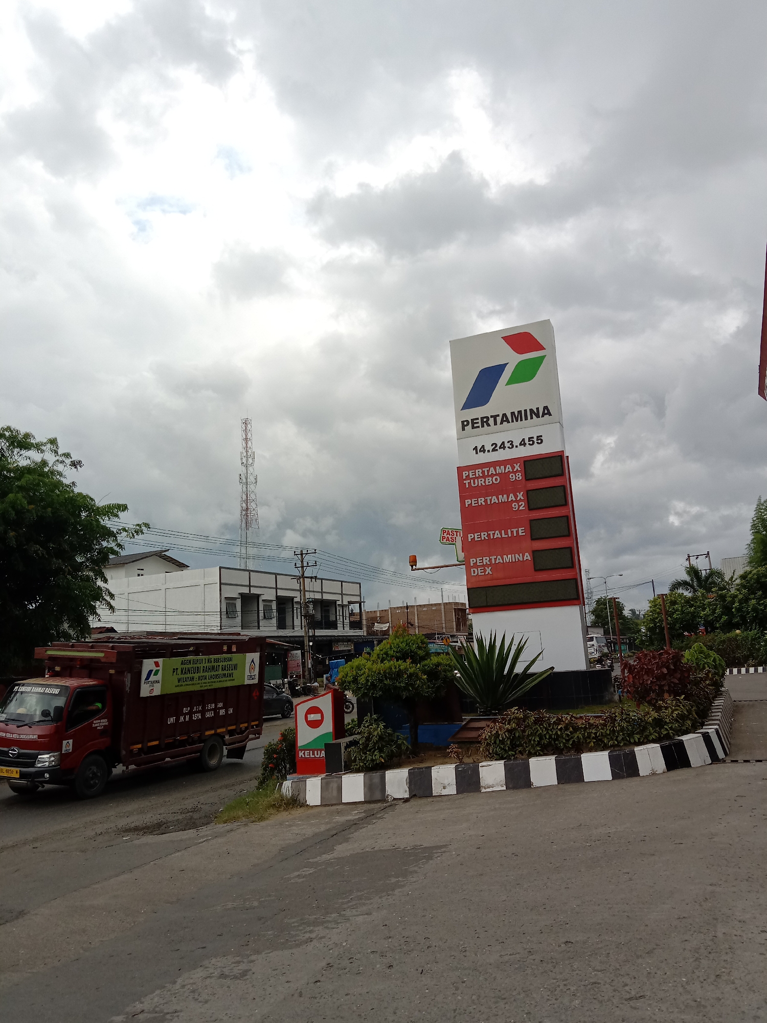 The image depicts a Pertamina gas station in Lhokseumawe, Indonesia. A large, vertical sign displays the company logo and prices for various fuel types: Pertamax Turbo 98, Pertamax 92, Pertalite, and Pertamina Dex. The prices themselves are not visible. In the foreground, there is a small roundabout with black and white striped curbing, planted with shrubbery. A red delivery truck with green Pertamina logos on the side is driving past the station. The truck has the following text on the side: "AGEN ELPIJI 3 KG BERSUBSIDI, PT. KANEUBI RAHMAT RASEUKI WILAYAH: KOTA LHOKSEUMAWE". A sign that reads "KELUA" is visible near the roundabout. The sky is overcast and gray. In the background, buildings and a cellular tower are visible.