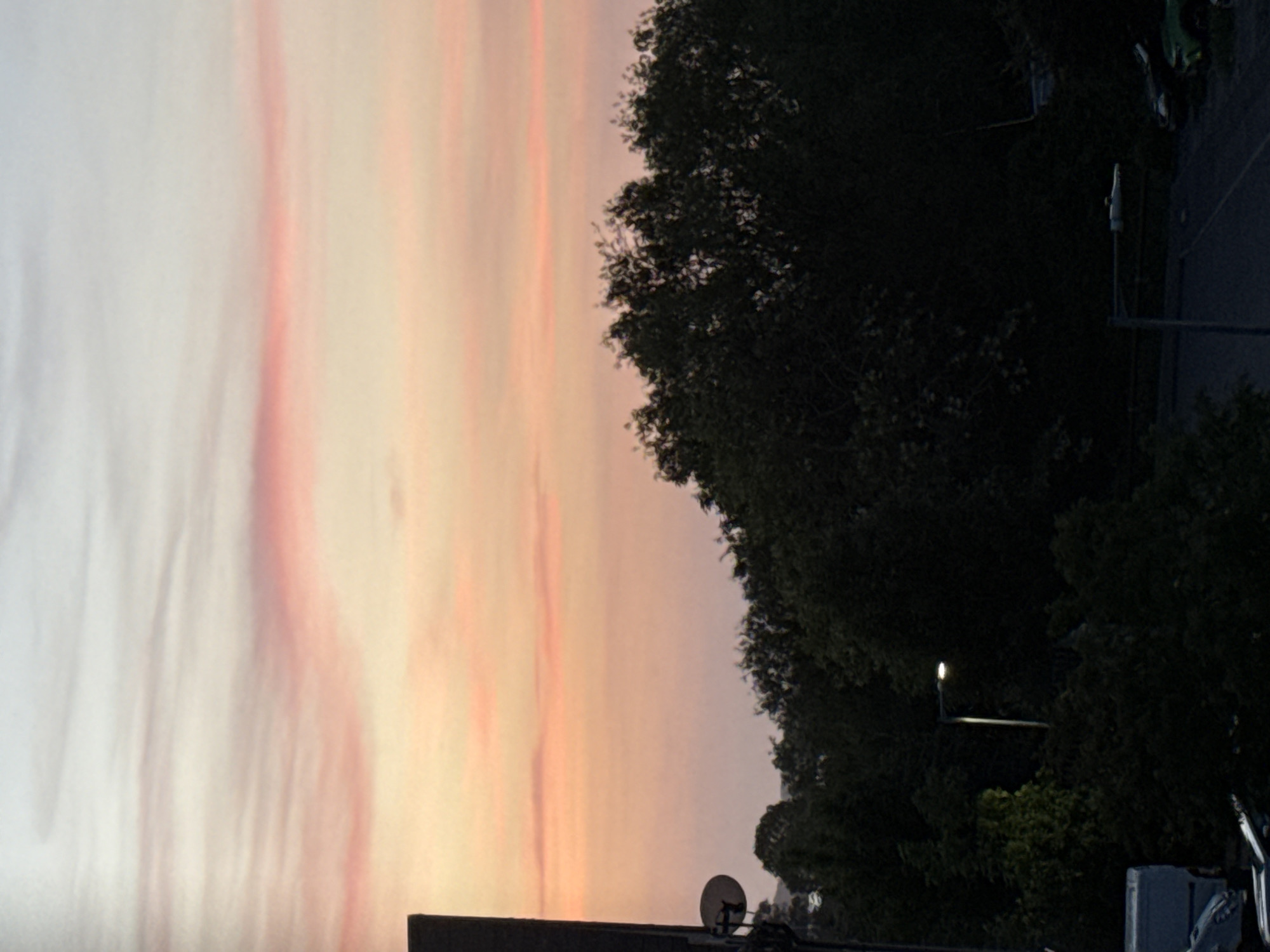 The image depicts a sunset in Great Yarmouth, United Kingdom. The sky is streaked with pink and white clouds, transitioning to orange and yellow nearer the horizon. To the right, dark green trees obscure a street. A street lamp and satellite dish are visible among the trees. A portion of a building is visible on the bottom center. There are parked scooters on the side of the road.