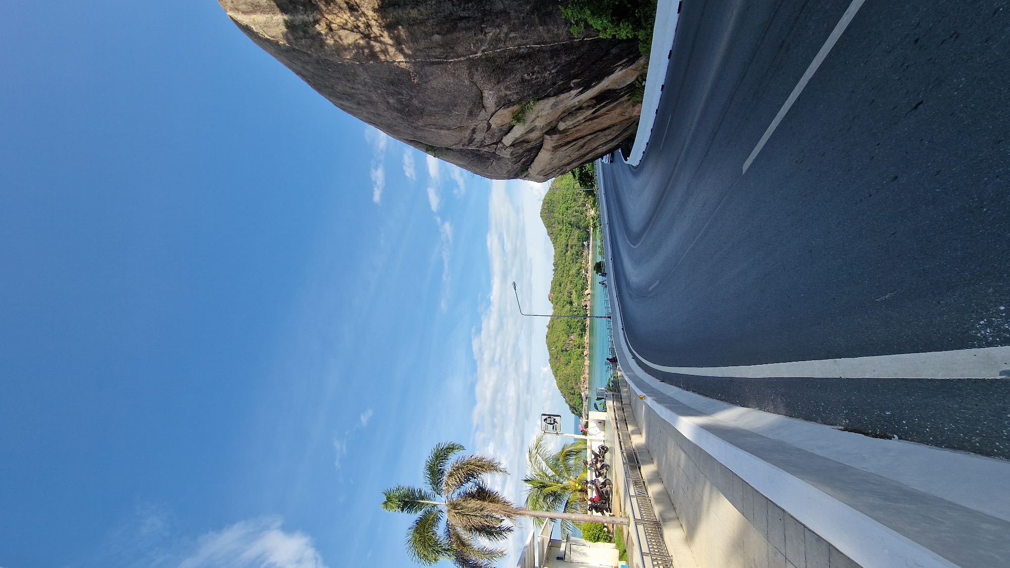 A winding asphalt road curves to the right, bordered by a white concrete barrier and a sidewalk with a metal railing. On the left, a palm tree stands beside a building with motorcycles parked in front. In the background, the turquoise ocean meets lush green hills under a bright blue sky with scattered clouds. A large, weathered boulder dominates the right side of the frame.