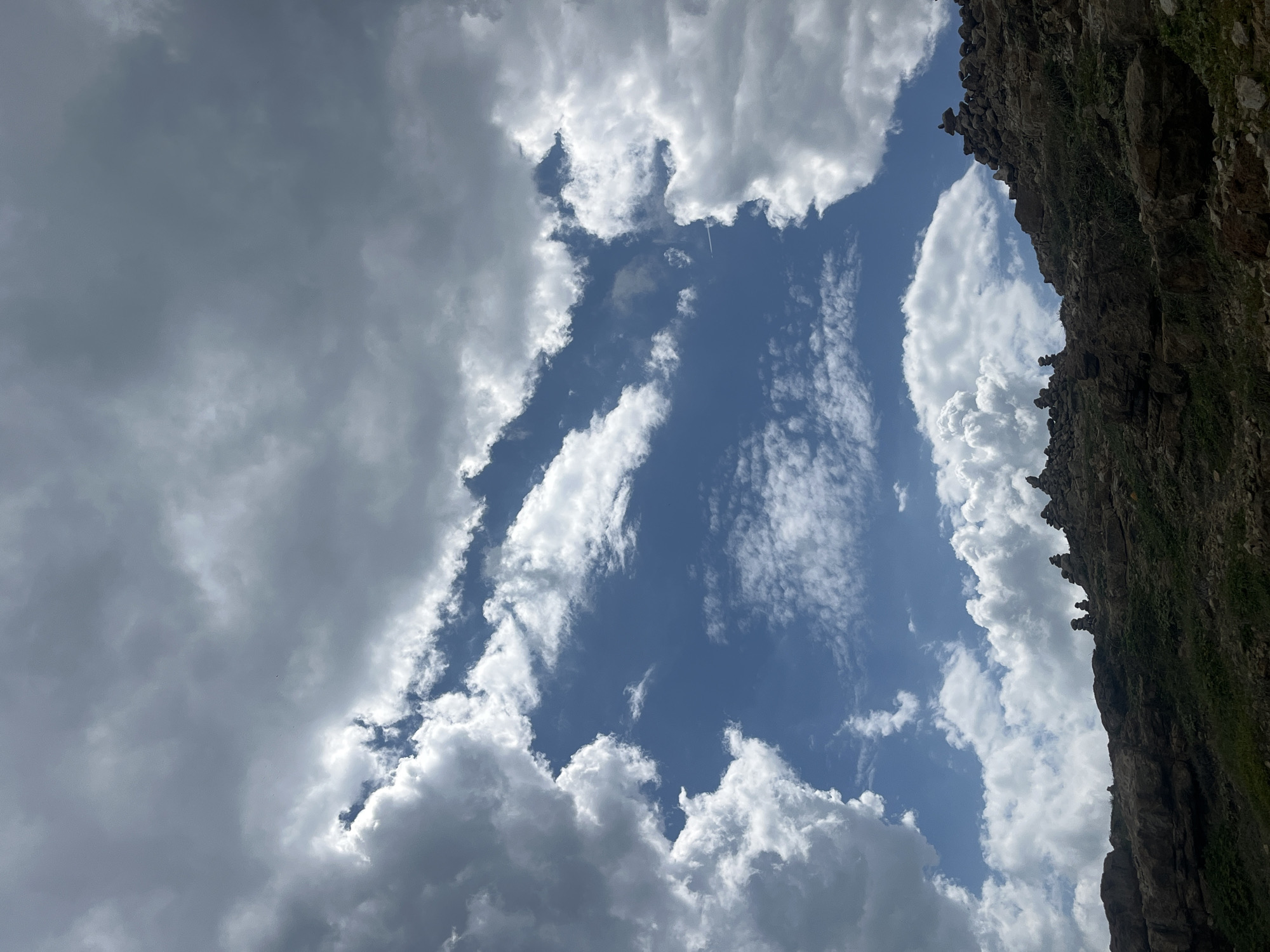 The image shows a rocky, mountainous landscape under a dramatic sky filled with clouds. The foreground is dominated by a rocky ridge with sparse vegetation. Several cairns, or stacks of rocks, are visible along the ridge line. The sky above is a mix of bright blue patches and large, billowing clouds, some of which are dark and ominous while others are bright white, suggesting sunlight filtering through. The overall impression is one of a rugged, natural environment. There is no indication of the city of Mui Ne, Vietnam, in this image.