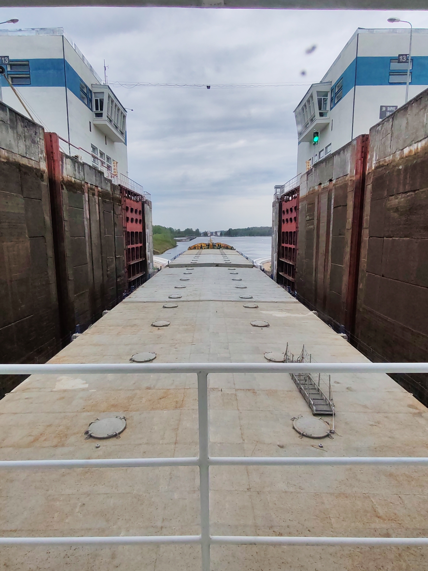A view from a barge traveling through a canal lock. The canal walls are high and constructed of weathered concrete. On either side of the canal, there are white control towers with blue accents and the number "13" visible on both. On the right control tower, there is a green traffic light visible. The barge's deck, made of concrete, features multiple circular access points. A white railing is visible in the foreground. The sky is overcast and gray. The location is Unknown, Unknown.