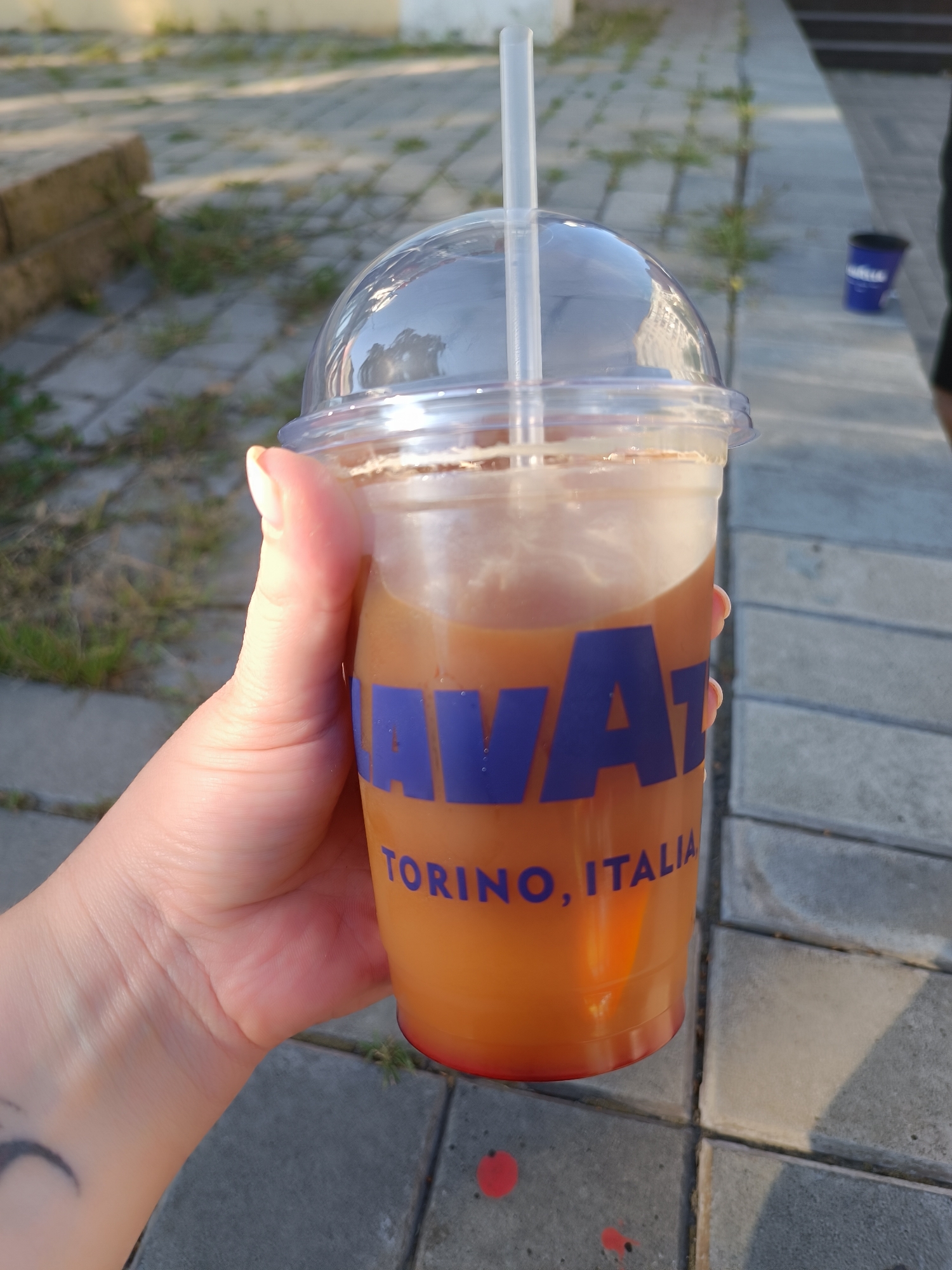 A hand holds a clear plastic cup filled with a light brown liquid, likely iced coffee, with a clear domed lid and a white straw. The cup has blue lettering that reads "LAVAZZA TORINO, ITALIA". The background is an outdoor scene with a paved walkway, some grass, and a stone structure. In the distance, a blue cup is visible on the ground. The lighting suggests it is daytime, possibly late afternoon.