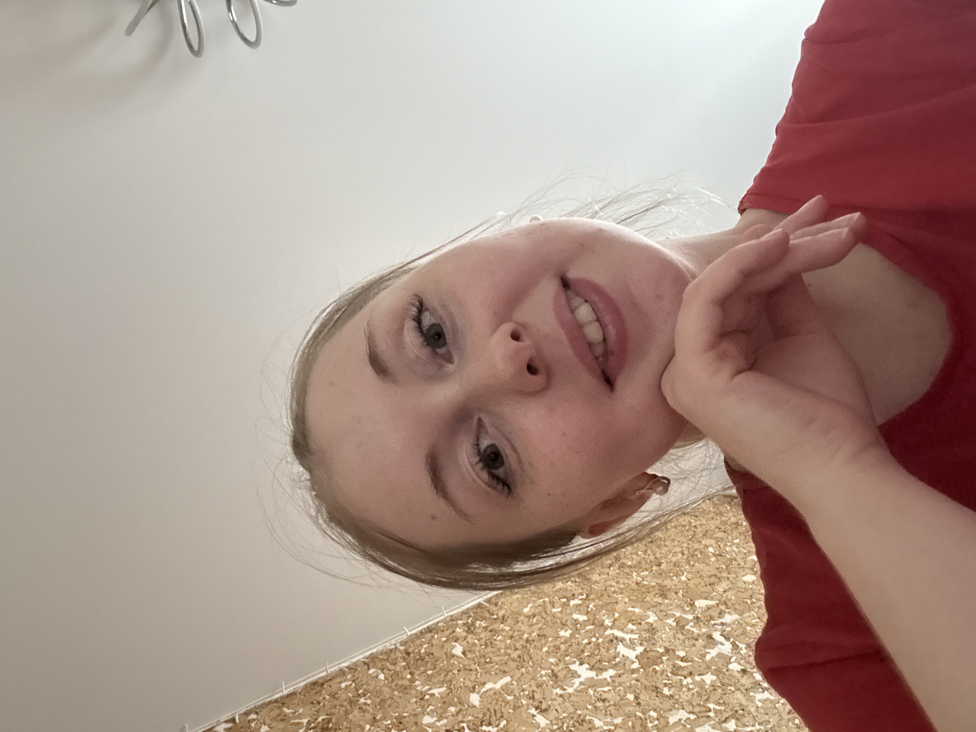 A low-angle selfie captures a young person looking down into the camera. The subject, likely female, has fair skin, light-colored hair, and light eyes. Her head is tilted to the right, and she wears a subtle, pleasant smile, revealing some teeth. Her right hand (viewer's left) is gently resting against her jawline/cheek. She is wearing a red top, visible on her right side.
The background is divided diagonally. The upper left portion is a plain, light-colored (likely white or off-white) ceiling or wall, with several metallic, spiral or ring-shaped objects visible in the top left corner, possibly part of a light fixture or decoration. The lower right portion of the background features a textured surface composed of irregular, light brown and white pieces, resembling cork board or a similar decorative material on a wall or floor. The lighting is bright and even, suggesting an indoor setting. There is no visible text in the image.