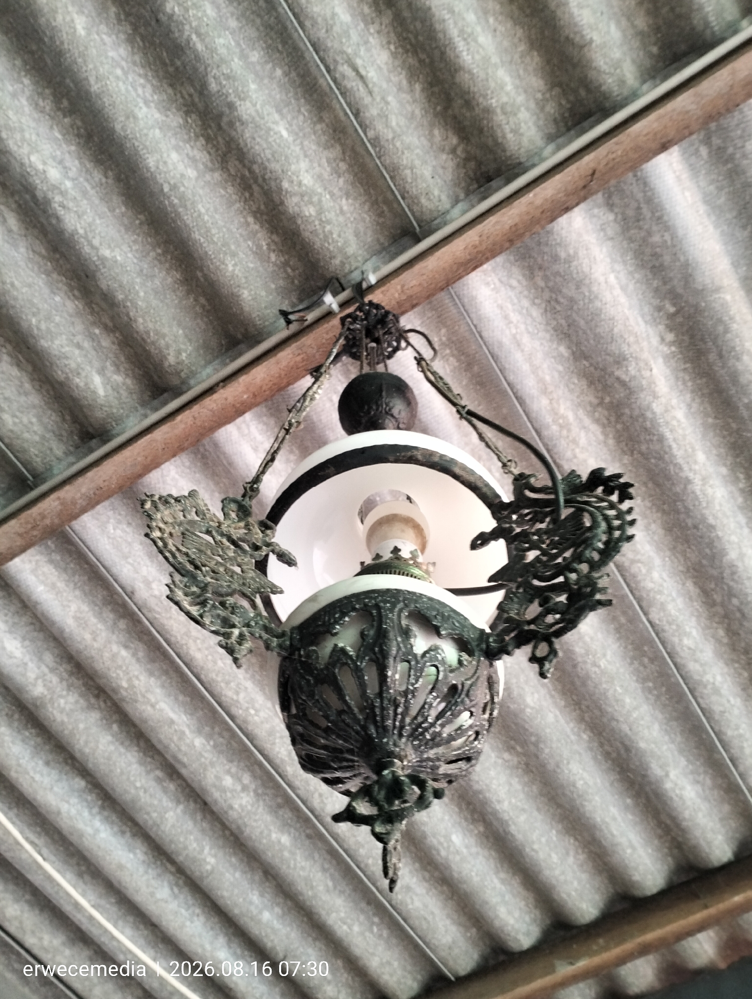 A low-angle shot captures an antique, ornate hanging lamp suspended from a corrugated roof structure. The lamp features intricate, dark metalwork resembling mythical creatures or dragons on either side, with a white bowl-shaped interior housing a lightbulb. The ceiling appears to be constructed from grey, ridged corrugated material, supported by a wooden beam. The lower left corner of the image displays a watermark: "erwecemedia | 2026.08.16 07:30". The scene is static and appears to be located outdoors or in an open-air structure, possibly in a residential or rustic setting in Imogiri, Indonesia.