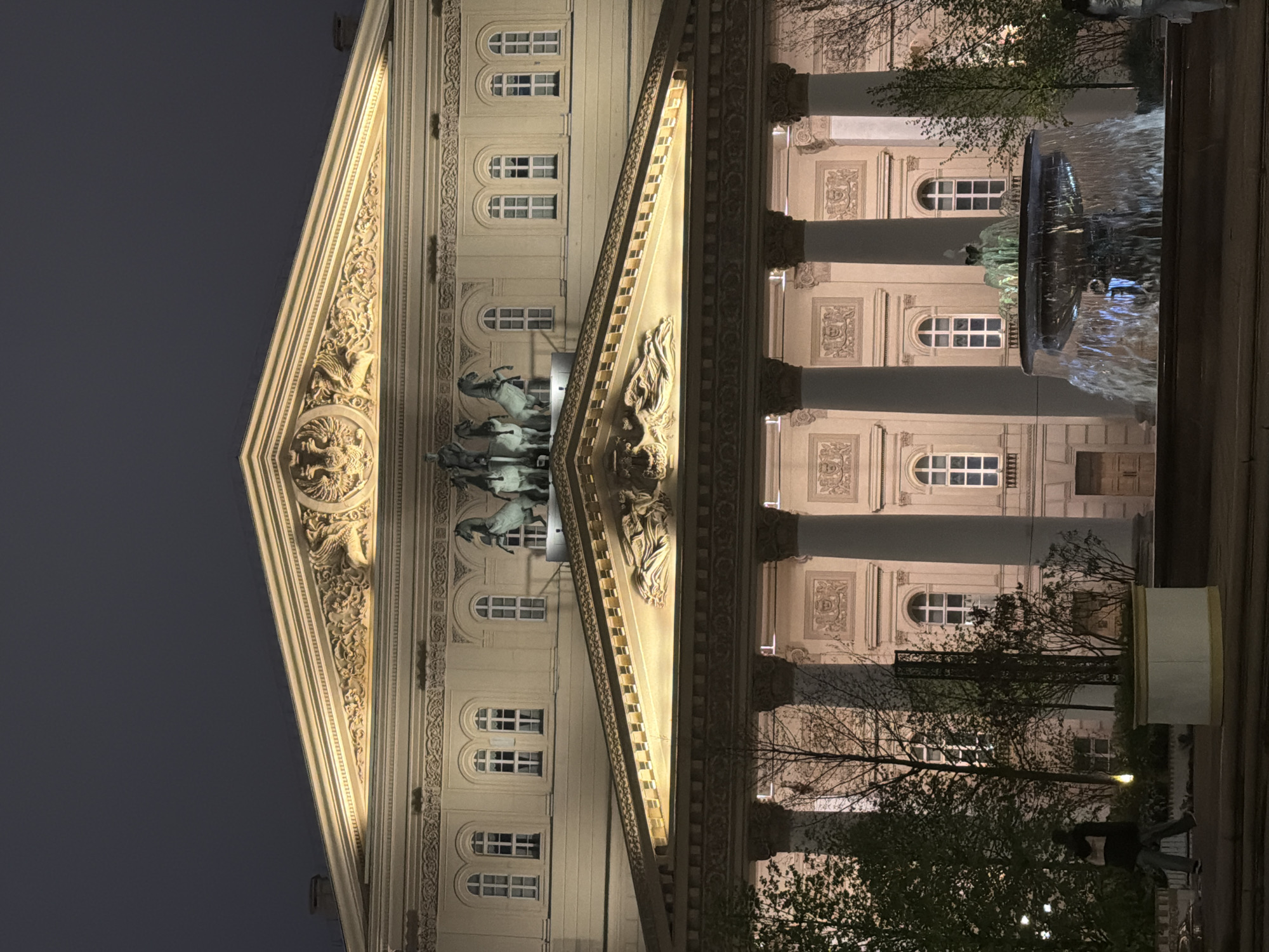 The image captures the Bolshoi Theatre in Moscow, Russia, at night. The facade of the building is illuminated, highlighting architectural details like the pediment, sculptures, columns, and windows. Above the main entrance, a statue of Apollo in his quadriga is visible. To the right of the theater, a fountain is gushing with water, partially obscuring the view of the building behind it. In the foreground, trees and silhouettes of people are present. The sky is dark.