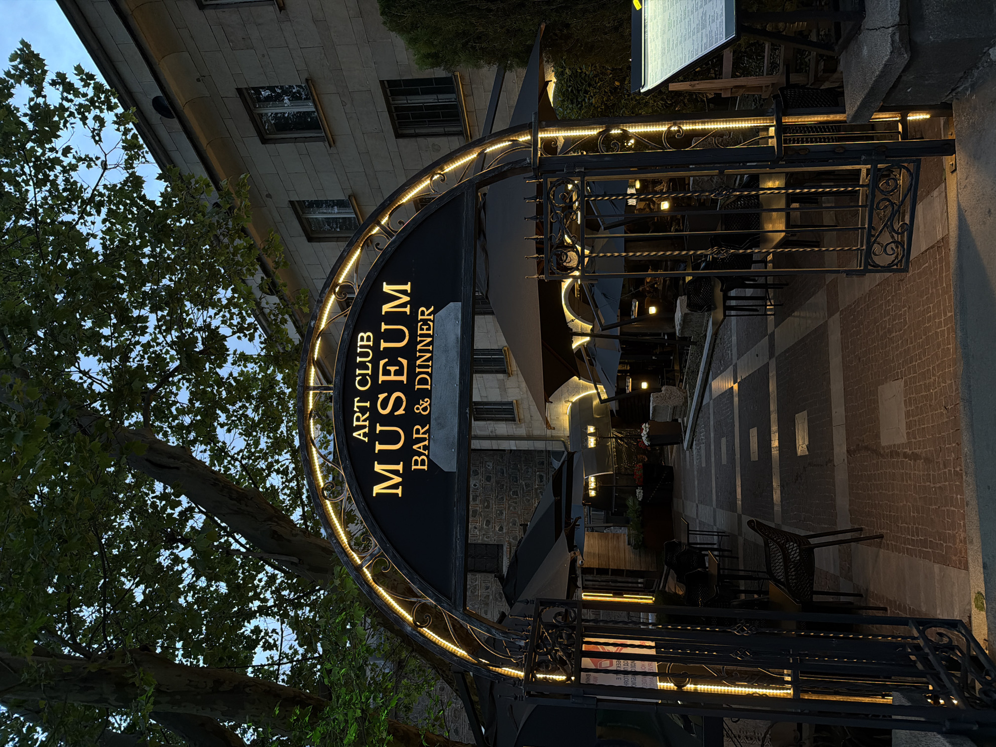 The image depicts the entrance to the "Art Club Museum Bar & Dinner" in Sofia, Bulgaria, as indicated by the sign. The entrance is framed by a black, ornate archway adorned with LED strip lights. Beyond the entrance, there is an outdoor dining area with tables and chairs under umbrellas, suggesting a restaurant or bar setting. The architectural style of the building in the background is European, with multiple windows and a stone facade. A tree with dense foliage is visible to the left of the archway. The scene is dimly lit, likely captured in the evening.