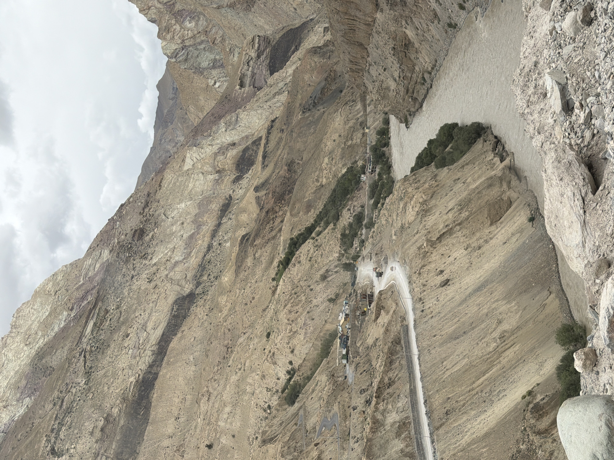 A wide-angle, high-angle shot captures a panoramic view of a mountainous landscape near Yulchung, India. A muddy, fast-flowing river cuts through a deep canyon. On the left side of the canyon, a winding road is carved into the steep, tan-colored mountainside. Several buildings, possibly a small village or settlement, are visible along the road. Trucks and cars are present on the road. The mountains are rugged and appear to be composed of sedimentary rock layers, with varying shades of tan, brown, and some greyish-purple hues. The sky is overcast with grey clouds. The overall impression is one of a remote and arid mountainous region.