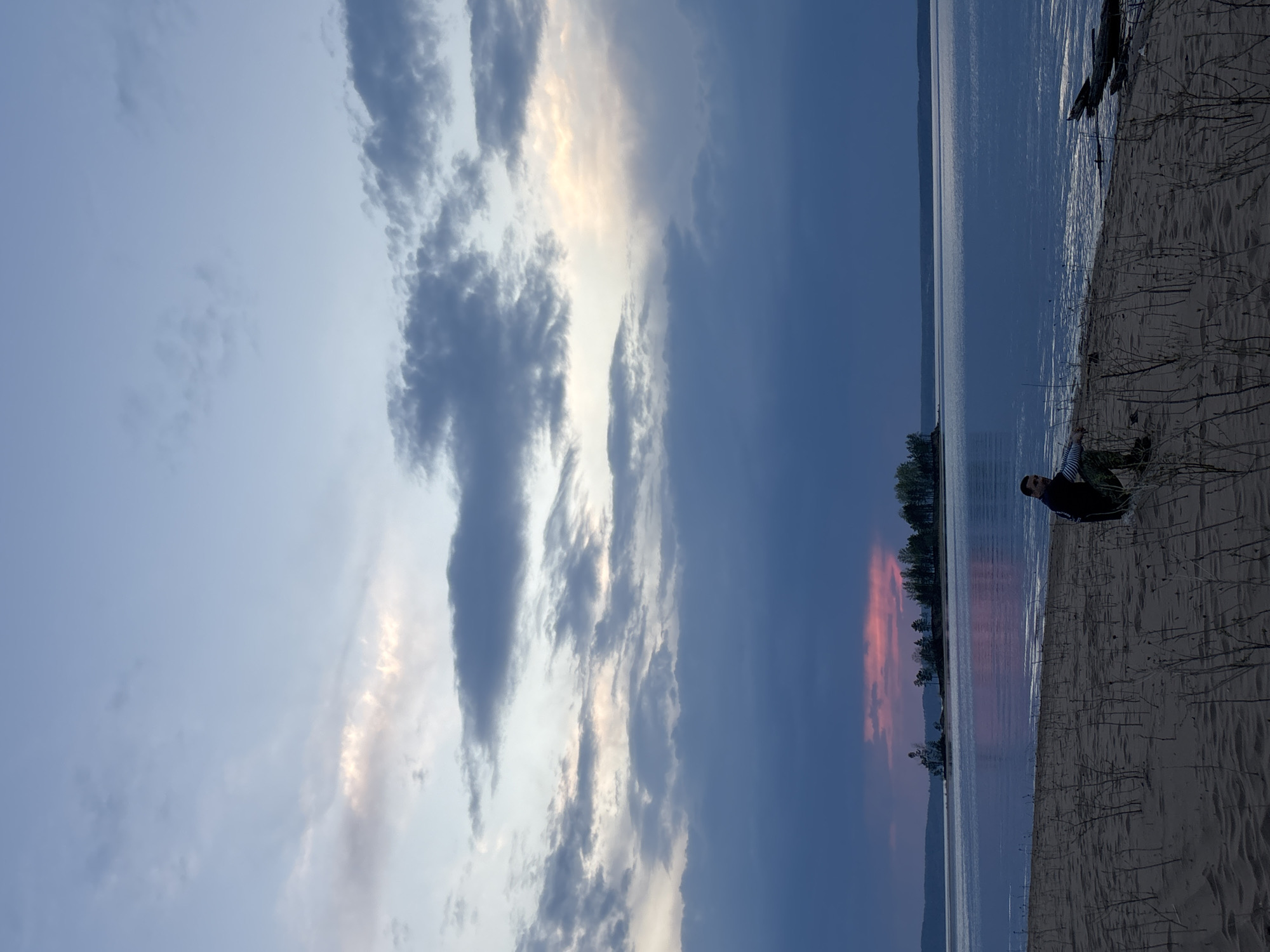 The image shows a shoreline scene at dusk in Vekoshinka, Russia. The sky is filled with clouds, transitioning from lighter shades to darker blue and grey. There's a band of orange and pink reflected on the water near the treeline on the opposite shore. The shoreline is sandy and covered with vegetation. A person sits near the edge of the water. A log is present on the sand.