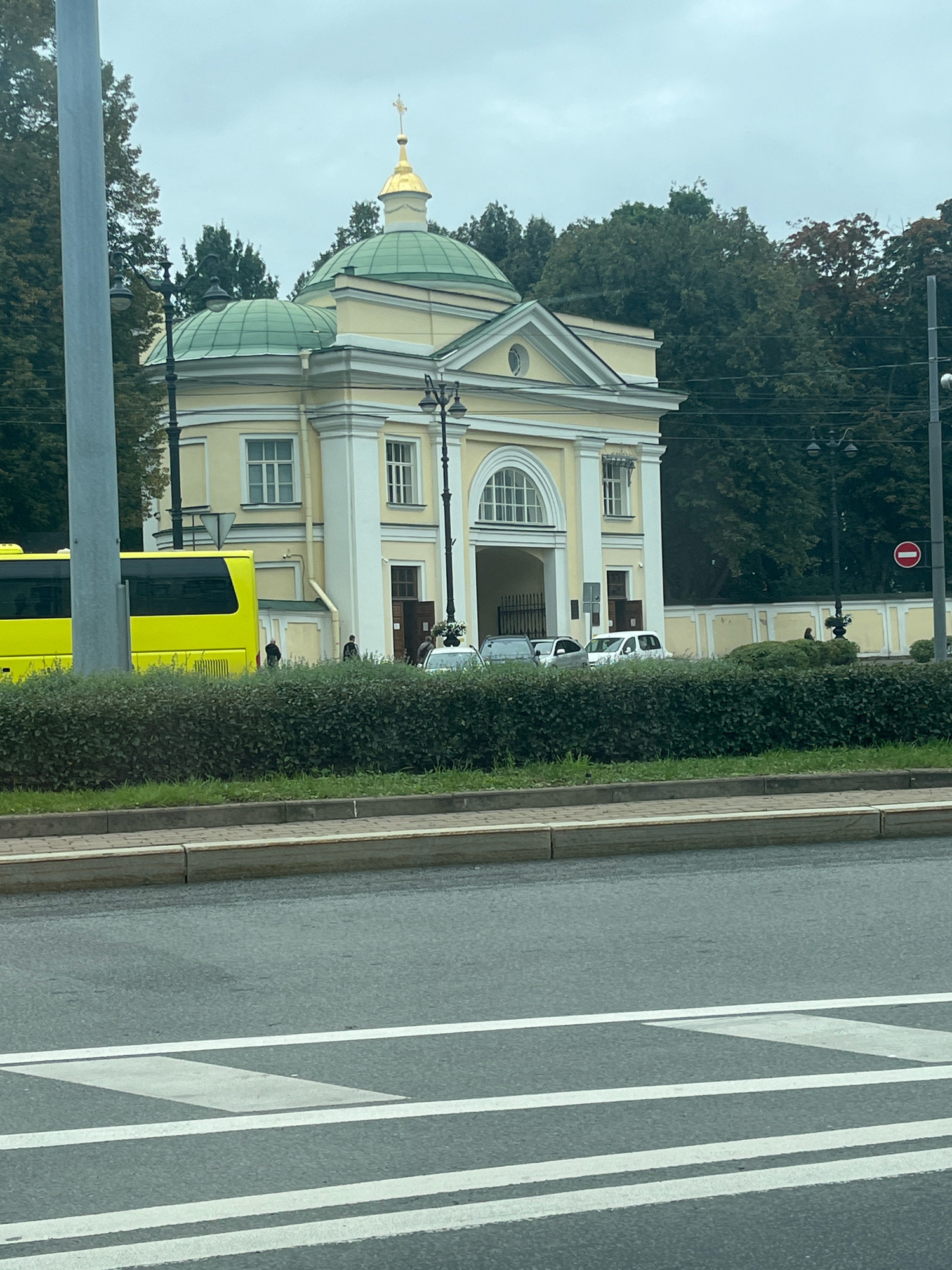 The image shows a light yellow building with a green dome and a golden cross on top, possibly a church, located in Saint Petersburg, Russia. The building has classical architectural elements, including columns and arched windows. In front of the building is a neatly trimmed hedge, and a street with white markings is visible in the foreground. A yellow bus is parked to the left of the building, and several cars are parked in front of the entrance. The sky is overcast, suggesting a cloudy day.