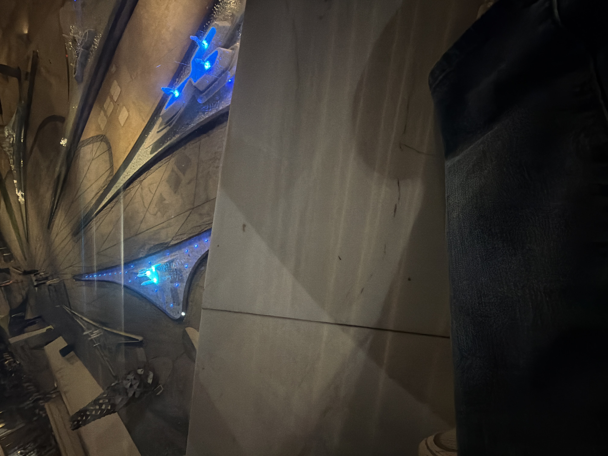 The image shows a dimly lit indoor scene with a focus on a model of a structure or landscape. The model features geometric shapes and pathways, some of which are illuminated with blue LED lights. In the foreground, a portion of a person's legs, clad in dark denim jeans, is visible. The background depicts a dark, textured surface, possibly a wall or floor. The overall impression is that of an artistic installation or an architectural model viewed from above, possibly in a public space. The mention of Nicosia, Cyprus, suggests the location of this scene.