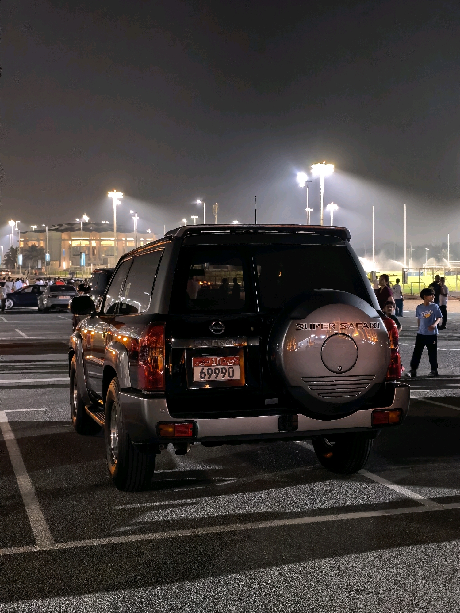 In a dimly lit outdoor parking lot at night, likely in Abu Dhabi, United Arab Emirates, a black Nissan Patrol SUV is parked. The vehicle is facing away from the camera, and its rear is prominently displayed. A spare tire cover with "SUPER SAFARI" inscribed on it is mounted on the back. A license plate with Arabic script and the number "69990" is visible. In the background, several tall floodlights illuminate a grassy area, possibly a sports field. People are scattered around the area, some standing and observing, while others are walking. In the distance, other vehicles, including a dark blue car, are parked. The scene appears to be outdoors, possibly at an event or a public gathering.