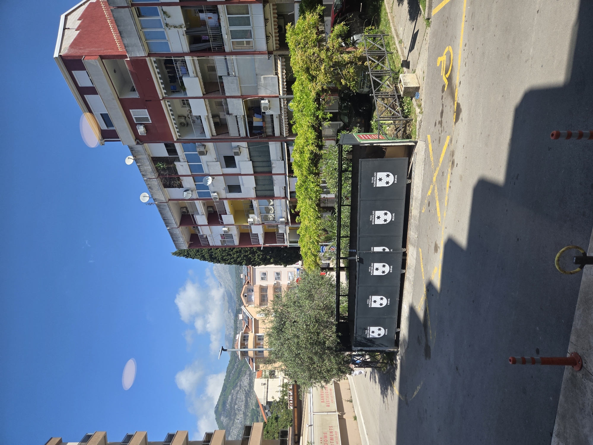 The image depicts a street scene in Budva, Montenegro, with a focus on urban elements. In the foreground, a row of black public waste containers is visible, adorned with the logo of "D.O.O. Komunalno Budva". Behind the containers is a tree and a section of Banbu Restaurant as well as some apartments for rent. The street itself has yellow painted markings, notably the symbol for disabled parking, and a red and white traffic cone. The surrounding environment includes apartment buildings with balconies and satellite dishes, a blue sky with light clouds, and a glimpse of mountains in the distance. The architecture appears modern with elements of classic Balkan design.
