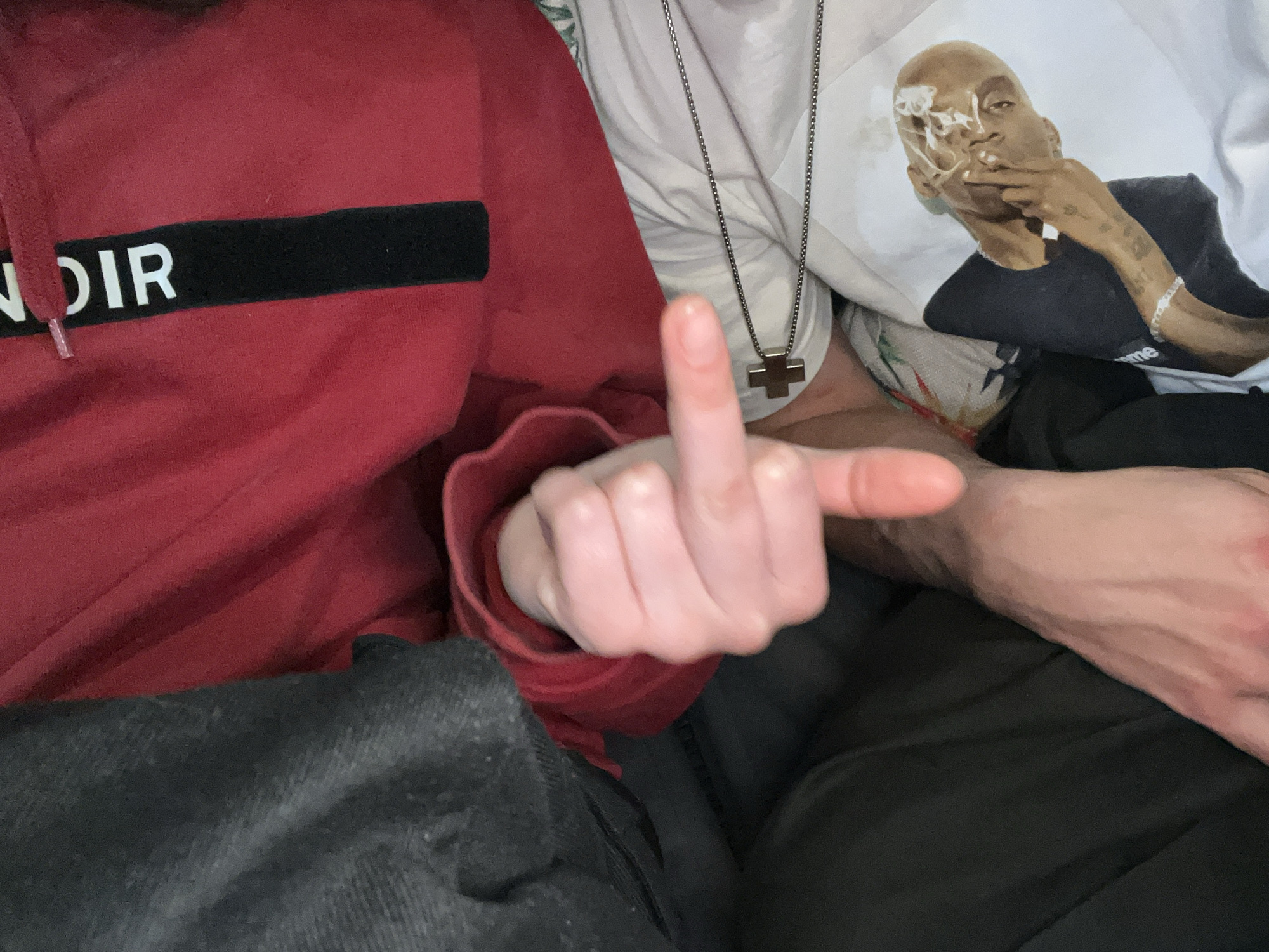The image shows a person wearing a red hoodie with the word "NOIR" visible on the front. Another person is wearing a white t-shirt with a portrait of a man smoking. A silver chain with a cross pendant hangs over the shirt. A hand with the middle finger extended is visible in the foreground. The background shows clothing, likely dark-colored pants. No information relates the image to Sochi, Russia.