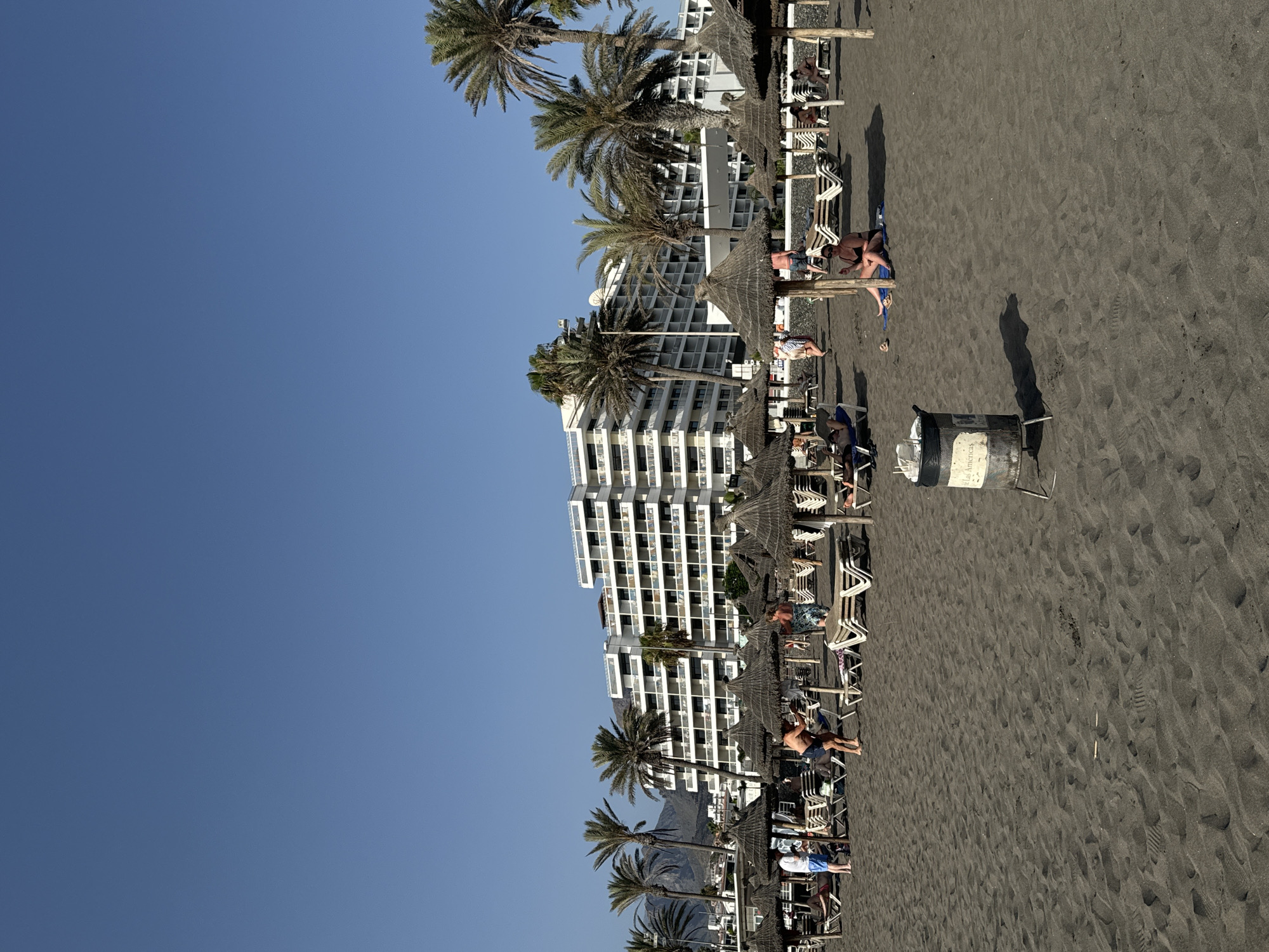 This image captures a sun-drenched beach scene in Costa Adeje, Spain. The foreground is dominated by dark, coarse sand, with footprints and scattered debris visible. Several thatched umbrellas and white lounge chairs are set up on the sand, some occupied by beachgoers. In the middle ground, a group of people are seen relaxing on lounge chairs or walking on the beach. Behind the beach setup, a large, multi-story white hotel with numerous balconies overlooks the scene. Palm trees are interspersed throughout the landscape, with mountains visible in the distant background under a clear blue sky. A metal trash can with a white lid and a label reading "Las Americas" stands prominently in the lower right section of the image, casting a shadow on the sand.