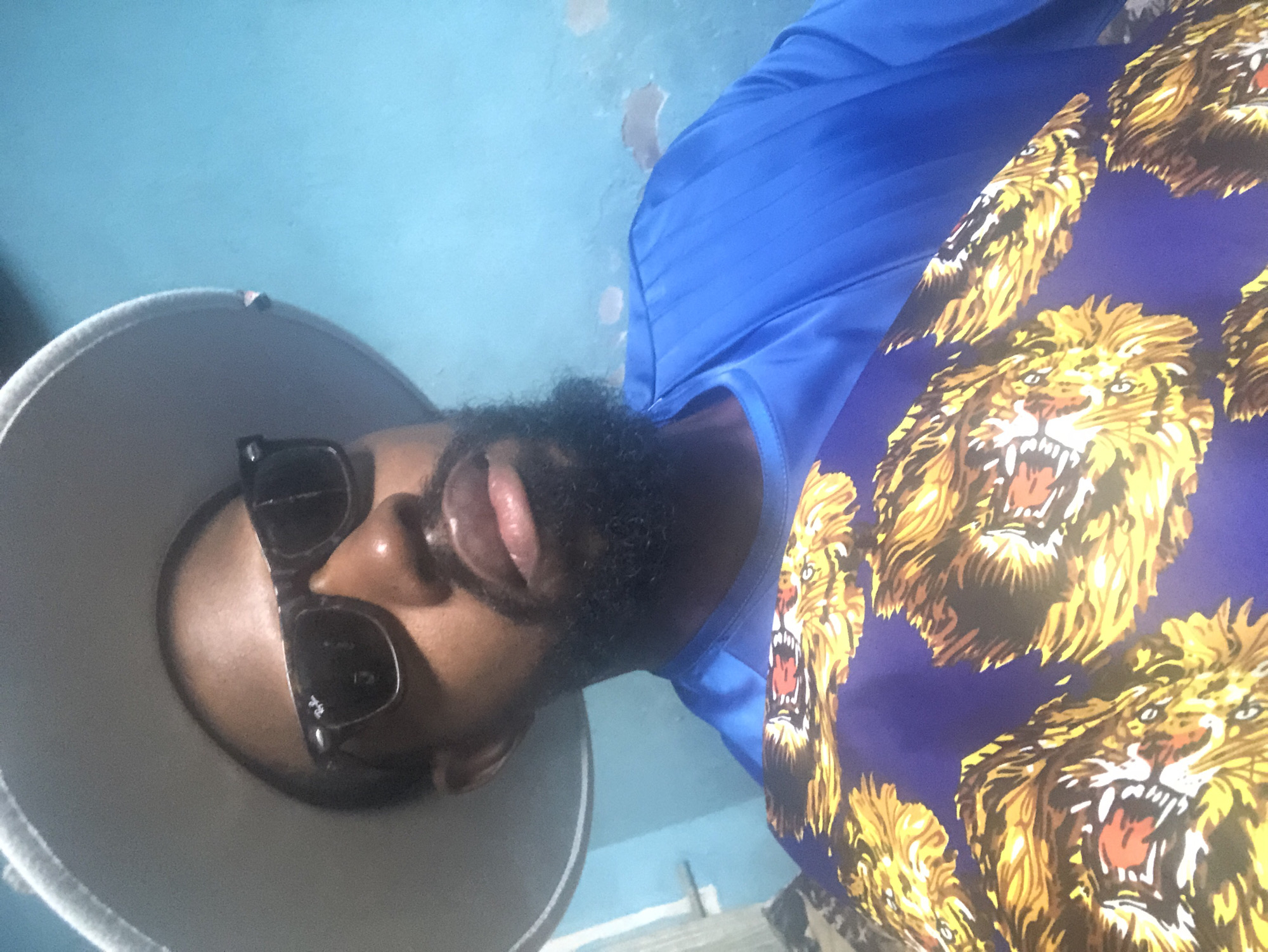A close-up, waist-up portrait of a Black man wearing a wide-brimmed light-colored hat, sunglasses, and a blue dashiki with a vibrant tiger print pattern. The man has a full beard and is looking towards the camera. The background is a bright, slightly textured light blue wall. The lighting suggests an indoor setting. The image quality is moderate.