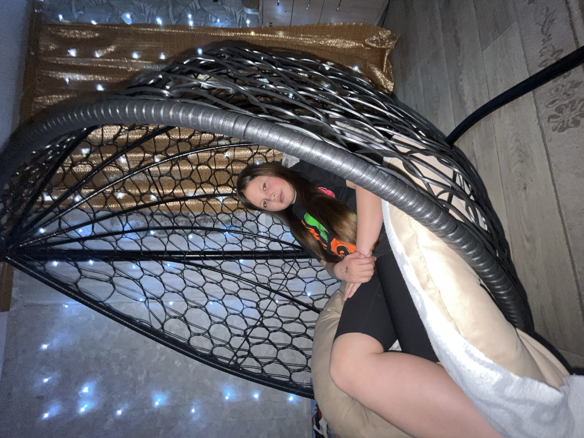A girl is sitting inside a black, egg-shaped, woven chair with a large beige cushion. The chair appears to be indoors, in a room illuminated by string lights. The wall behind the chair is covered with a gold-colored material. The floor is light-colored with a wood grain pattern. The image appears to be taken in Novopokrovskaya, Russia.
