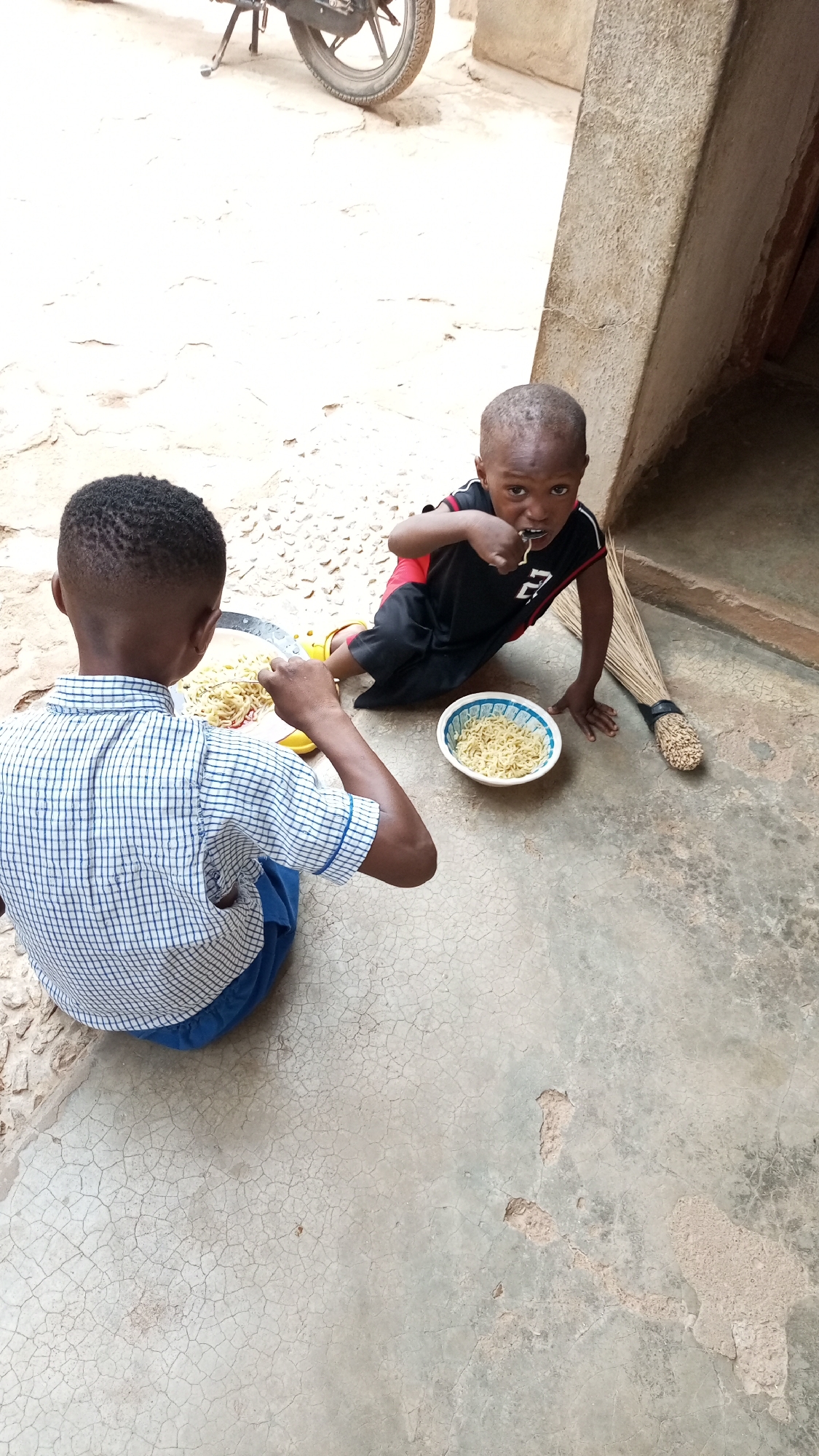 Two young children are eating noodles on a concrete surface outside. One child is wearing a blue and white checkered shirt and blue pants, facing away from the camera. The other child is wearing a black and red shirt and black shorts, looking directly at the camera while eating. A bowl of noodles sits beside him. A broom leans against a wall nearby. A motorcycle is partially visible in the upper left corner of the image. The location appears to be in a residential area with a concrete floor.