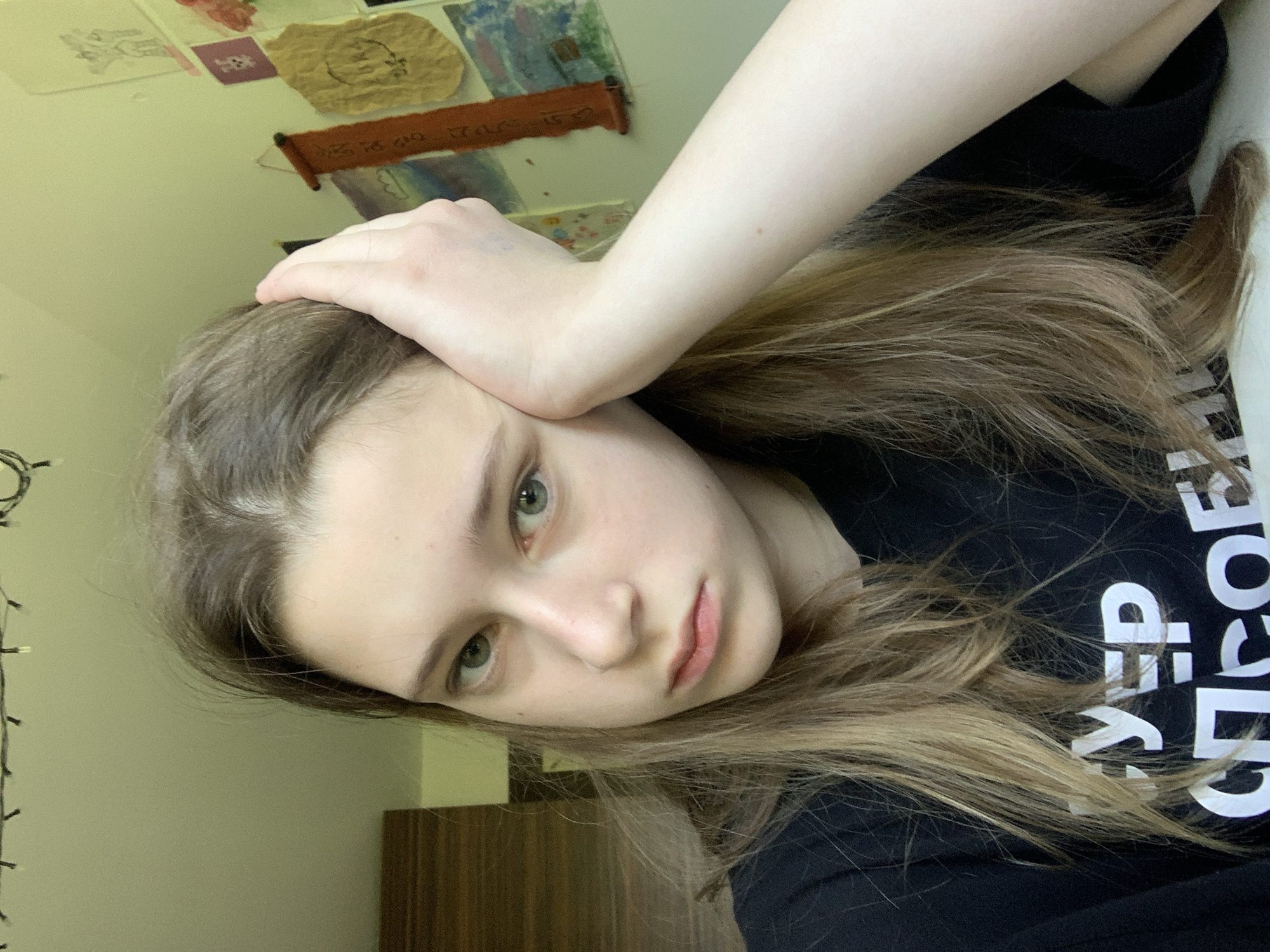 A fair-skinned young woman with light brown hair is angled to the left in the image. She has her right hand resting on her forehead. She is wearing a black shirt with white text. The background shows a wall with hanging decorations and string lights. The location cannot be determined from the image provided.