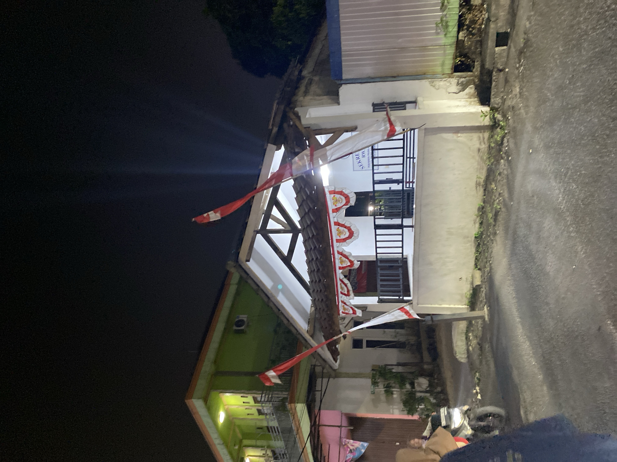 The image depicts a nighttime scene in what appears to be a residential or semi-commercial area in Tangerang, Indonesia. The primary focus is on a building with a white facade and a dark-tiled roof, decorated with red and white pennants. Two Indonesian flags are prominently displayed; one is a tall flag pole with a flag, and another is a tattered red and white flag draped across a pole. In front of the building, a black metal fence encloses an area. To the left of this building, another structure is visible, painted bright green on its upper level with balconies, and a pink section on its lower level.
A motorcycle is parked on the paved street in the foreground, and a person wearing a light brown headscarf is seated on it, looking towards the left. Another person, wearing dark clothing, is partially visible in the lower left corner, possibly walking or standing. The street itself is asphalt and appears wet, with some debris and vegetation growing along the edges. The overall lighting suggests artificial illumination, with some light beams emanating from the green building and from streetlights, creating a somewhat dark and atmospheric ambiance. The presence of Indonesian flags and pennants suggests a possible celebration or patriotic event.