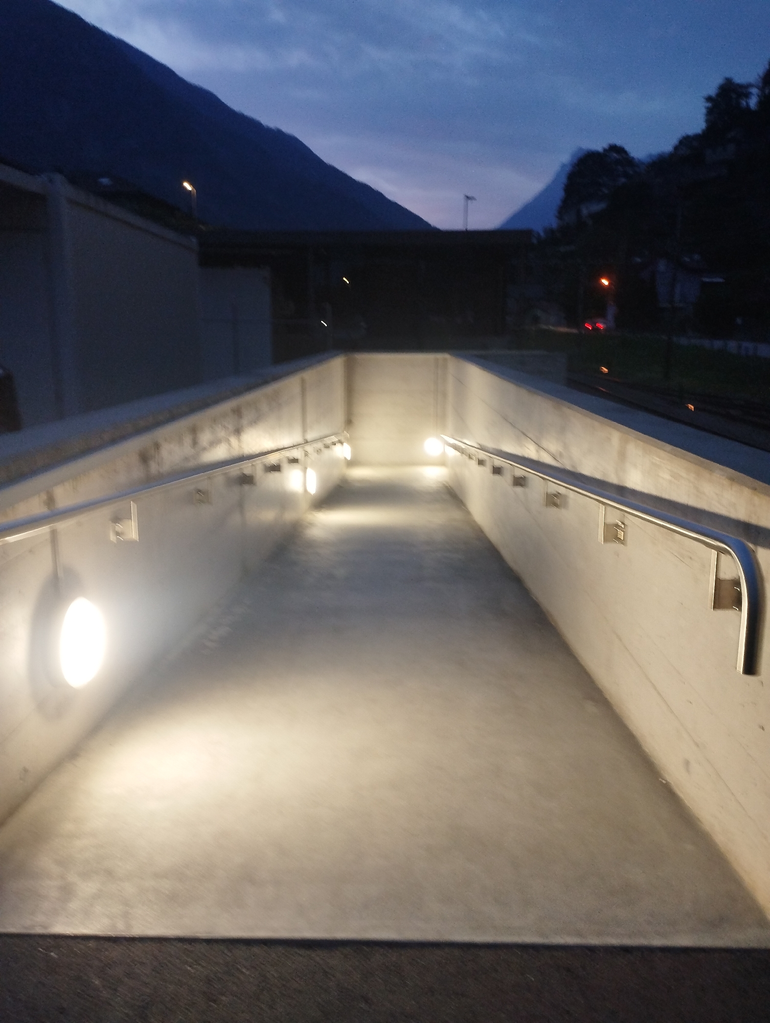 This is a vertical, eye-level, full shot of a dimly lit concrete pathway that slopes downwards. The pathway is bordered by concrete walls with a metal handrail running along the top. Several round, white lights are embedded in the left wall, illuminating the pathway. A few more lights are visible further down the pathway on the right wall. The sky above is a dark blue, with faint clouds visible. In the background, a dark mountain range rises against the sky, and the silhouettes of buildings and trees can be seen. The image appears to have been taken at dusk or at night.