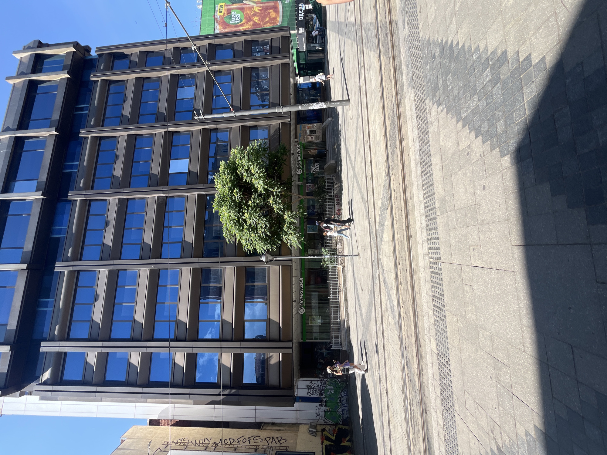 This is a vertical, eye-level shot of a modern, multi-story building with a dark facade and many blue-tinted windows. The building appears to be an office building or commercial space. In front of the building, there's a paved area with tram tracks running through it. A medium-sized tree with lush green foliage is positioned in front of the building, partially obscuring the lower floors.
Several people are visible in the scene. On the sidewalk to the right of the building, a few individuals are walking, including one person on a skateboard. Further down, a person is standing near the entrance of the building, and two more people are walking away from it. Graffiti is present on the lower part of the building's facade, near the ground level.
Above the building, a large billboard advertising "fuze tea" is visible. The sky is clear and blue, suggesting a sunny day. The lighting indicates it's likely daytime, possibly late morning or early afternoon, given the clear shadows cast on the ground. The presence of tram tracks and the Cyrillic text on the billboard and building suggest the location is likely in Eastern Europe, specifically Sofia, Bulgaria, as indicated in the prompt. The text visible includes "С банка дск" (which translates to "DSK Bank"), "NEW", "fuze tea", "ОДЕДН", and "ТУК СЕГА". The overall impression is an urban scene on a bright, clear day.