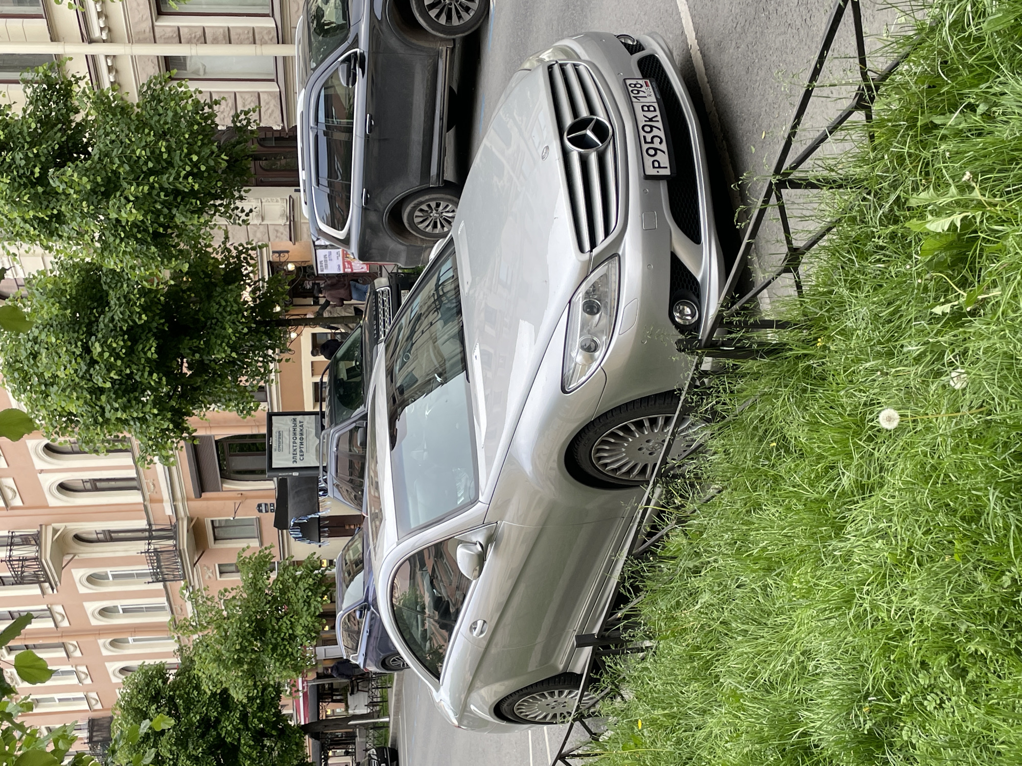The image depicts an urban street scene in Saint Petersburg, Russia, during the daytime. A silver Mercedes-Benz sedan is prominently parked in the foreground, angled towards the left. Its front grille, adorned with the Mercedes-Benz emblem, and the front passenger side are clearly visible. The car's Russian license plate, "P959KB 198 RUS," is affixed to the front bumper, with the "198" code identifying its registration in Saint Petersburg and the Leningrad Oblast. The vehicle features multi-spoke alloy wheels.
To the right of the Mercedes-Benz, a strip of overgrown green grass is visible, separated from the street by a low, dark metal fence. Behind the Mercedes-Benz, a dark grey SUV is also parked, with a partially visible "LAND ROVER" emblem on its rear.
The background is dominated by multi-story buildings with light-colored (beige or peach) facades, featuring numerous arched windows and decorative balconies, characteristic of historical European architecture common in Saint Petersburg. Green, leafy trees line the street, partially obscuring some of the buildings. On the sidewalk, a white sign with black Cyrillic text is visible; the most readable part states "ЭЛЕКТРОННЫЙ СЕРТИФИКАТ" (Electronic Certificate), with "ОТКРЫТАЯ" (Open) possibly above it.
The lighting is soft and natural, suggesting an overcast day or a period of day without direct harsh sunlight. The scene is calm, portraying a typical city street with parked vehicles and established urban greenery. No individuals or specific human activities are clearly discernible in the frame.