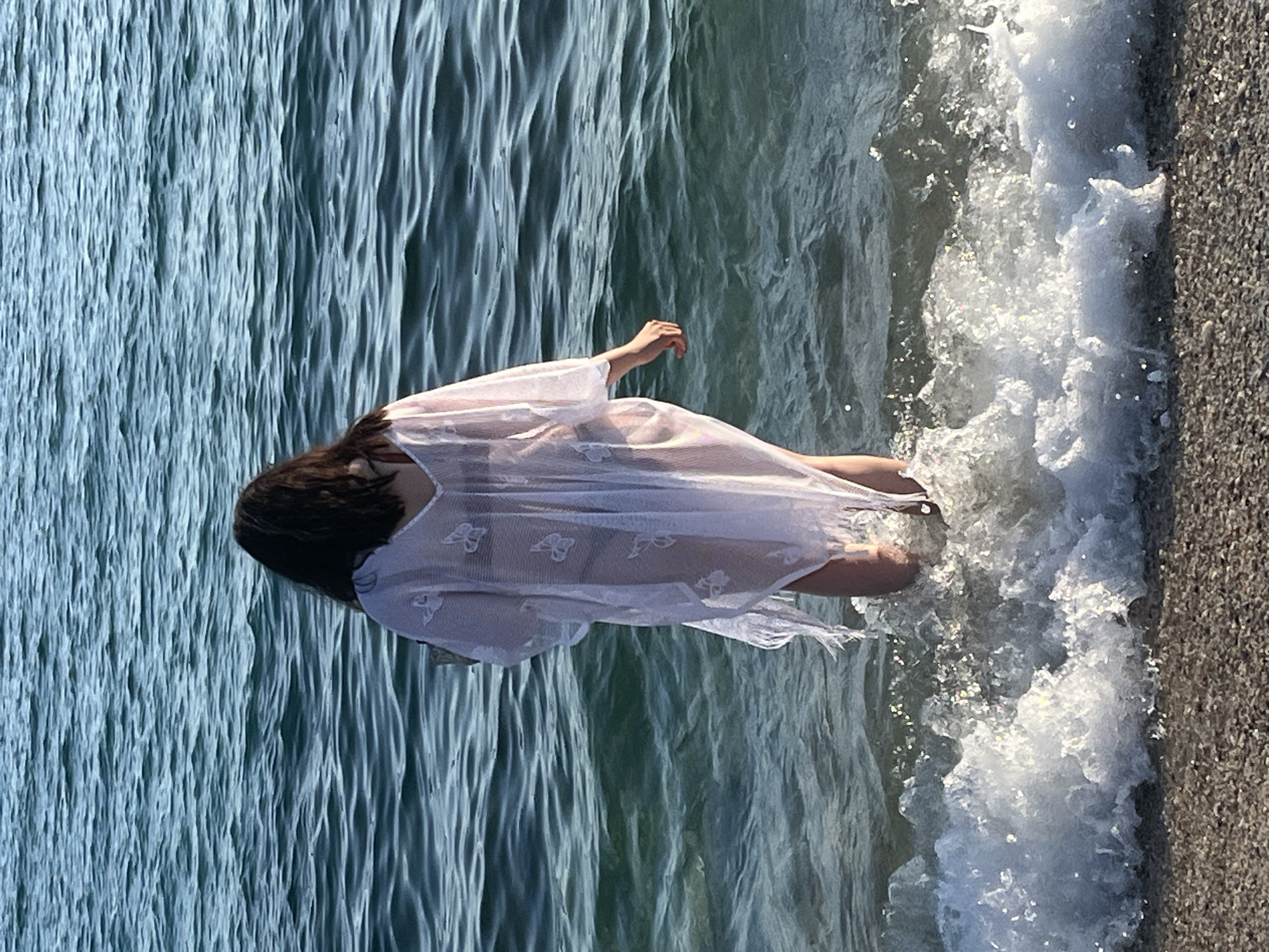 A woman wearing a white, sheer dress stands in the surf on a beach in Alanya, Turkey. The waves are foamy as they break on the shore. The water appears to be turquoise. The beach is made of small grey pebbles.