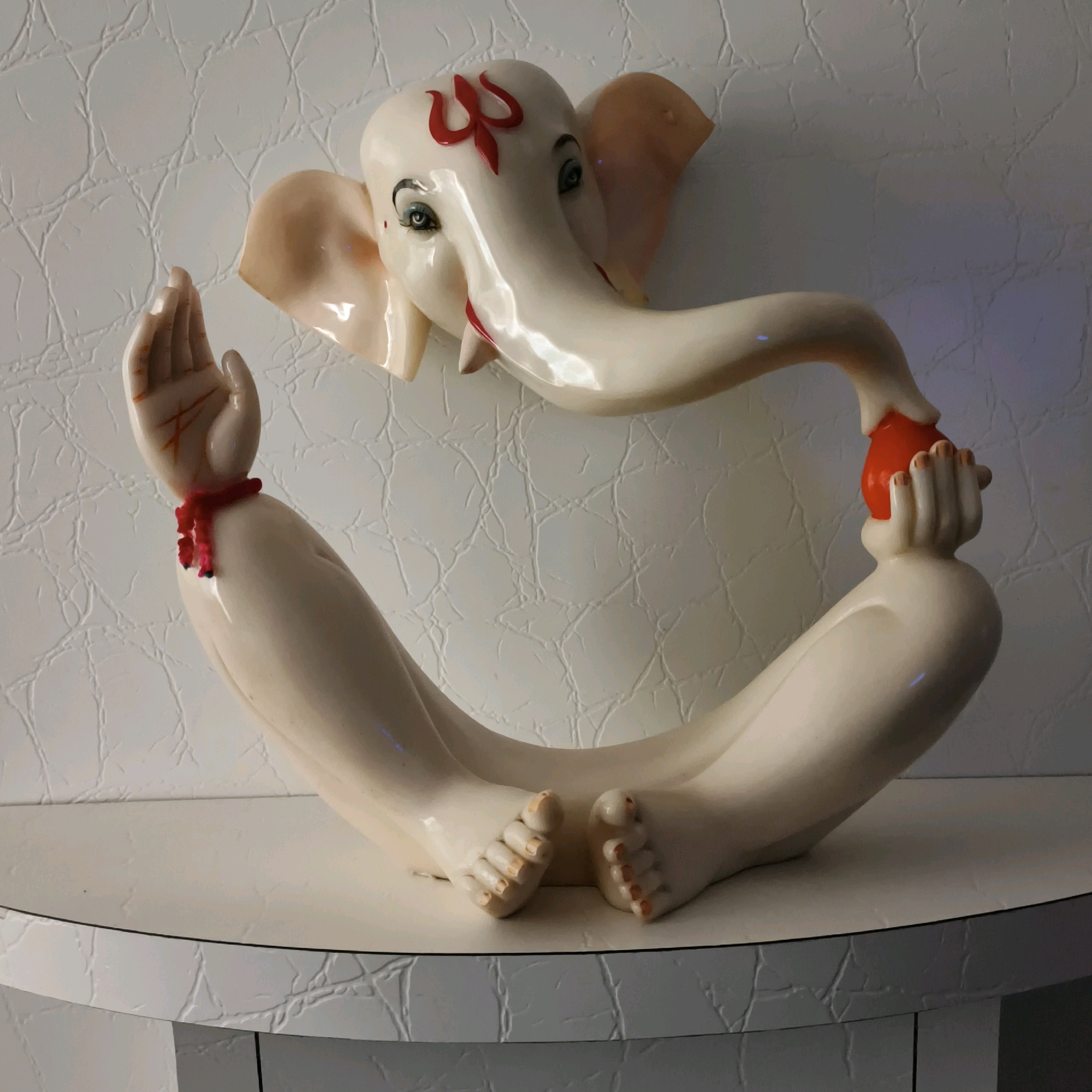 An artistic sculpture of Lord Ganesha, made of ceramic, is displayed on a white table against a textured white wall. The sculpture depicts Ganesha in a seated position with his legs crossed. His head is tilted forward, and his trunk is curved to hold a small, red, round object, possibly a fruit or sweet, in his left hand. His right hand is raised with the palm open, and a red string is tied around his wrist. Ganesha's forehead is adorned with a red "Trishul" symbol. The sculpture exhibits a glossy finish, and subtle lighting creates highlights and shadows. The background wall has a cracked, embossed pattern. The table has a rounded edge and a dark line detail. The overall scene is static and appears to be an indoor display. There is no indication of its specific location within Mumbai, India.