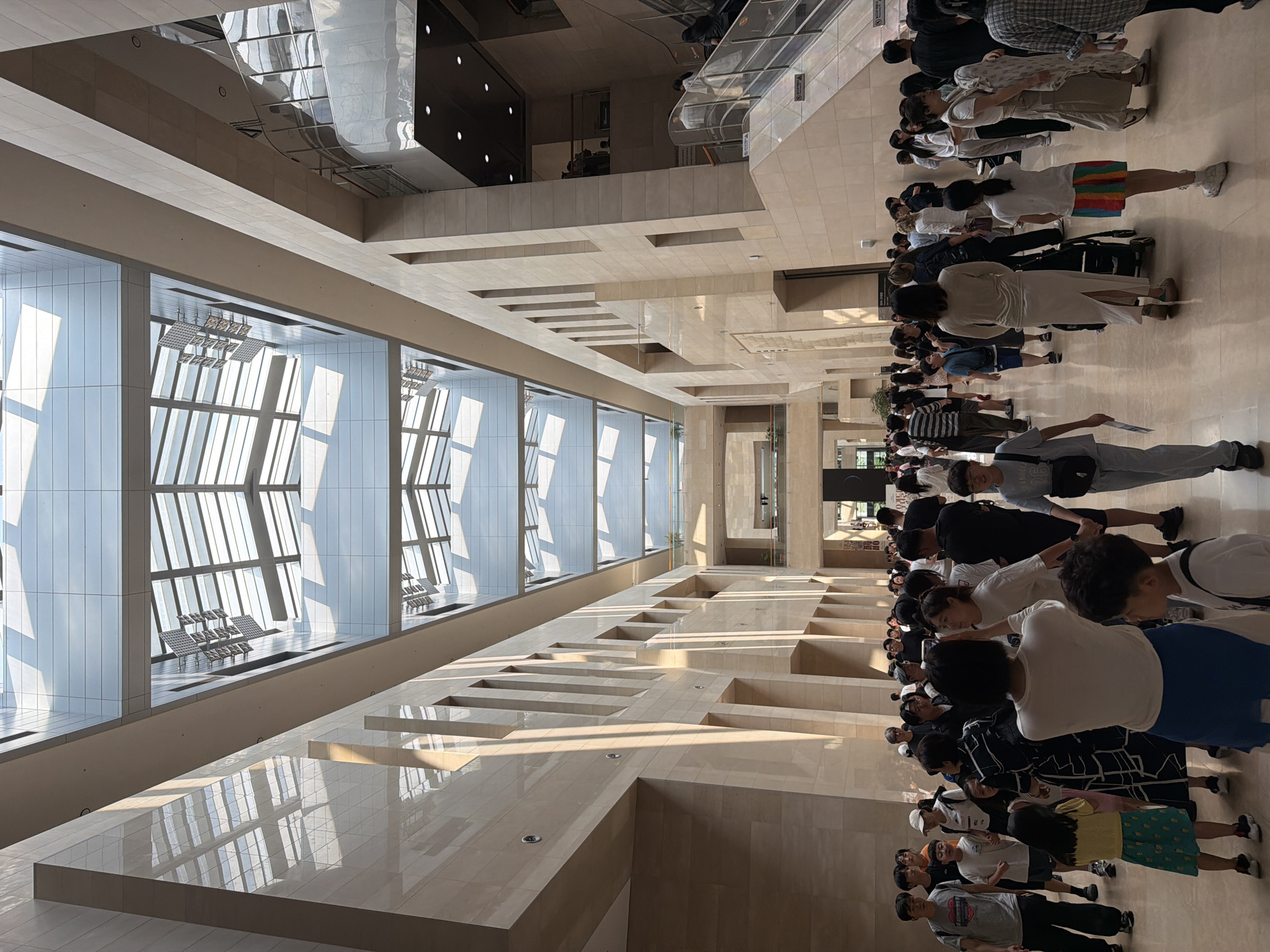 The image shows a wide, sunlit atrium with a high, modern ceiling consisting of glass panels and structural beams. The atrium is filled with people, suggesting it is a public space such as a museum, gallery, or convention center. The architectural style is contemporary, with smooth, light-colored walls and floors. Sunlight streams through the glass ceiling, creating geometric patterns of light and shadow on the floor and walls. Several groups of people are visible, some walking, some standing and talking, and some appearing to be taking photographs. The presence of stairs and escalators on the right side of the image indicates multiple levels within the building. Based on the architectural style and the presence of many visitors, this scene is likely located in a major cultural institution in Seoul, South Korea.