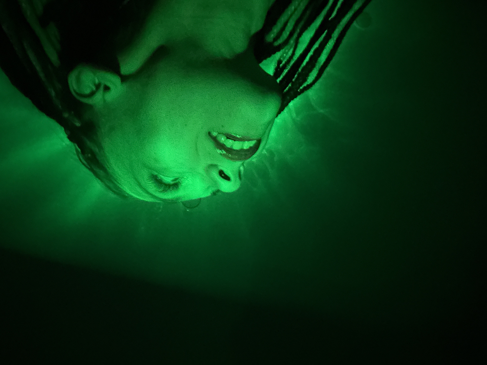 A close-up, low-angle shot shows a person's face and part of their head, seemingly underwater, illuminated with green light. The person is facing downward with their head tilted back. Their mouth is slightly open, revealing their teeth. A single drop of water is visible beneath the nostril. The person has dark braids framing their face. The background is dark green, fading into black. The scene appears to be set underwater in a location with artificial green lighting, but it is impossible to verify its location in Rusanovo, Russia, based solely on the image.