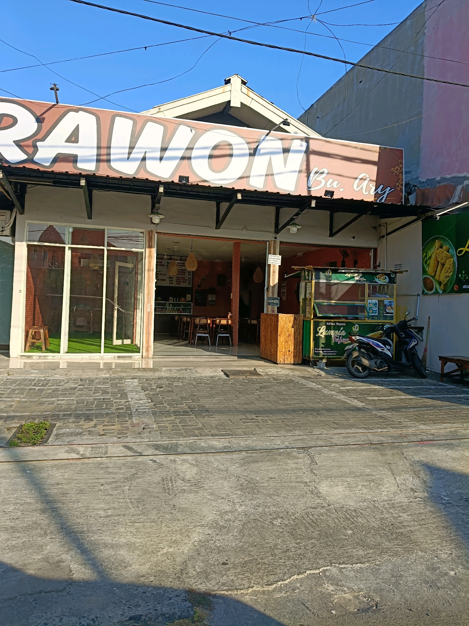 The image shows the exterior of a restaurant in Surabaya, Indonesia, named "Rawon Bu Ary." The building has a prominent sign with large white letters spelling out "RAWON." To the right of the sign, smaller cursive text reads "Bu. Ary." The restaurant features a glass-fronted entrance on the left and an open doorway on the right, revealing seating areas inside. Outside, a food cart with green and yellow accents is parked in front of the entrance. A motorcycle is parked next to the food cart. The ground in front of the restaurant is paved with bricks and concrete. The sky is clear and blue, suggesting it is daytime.