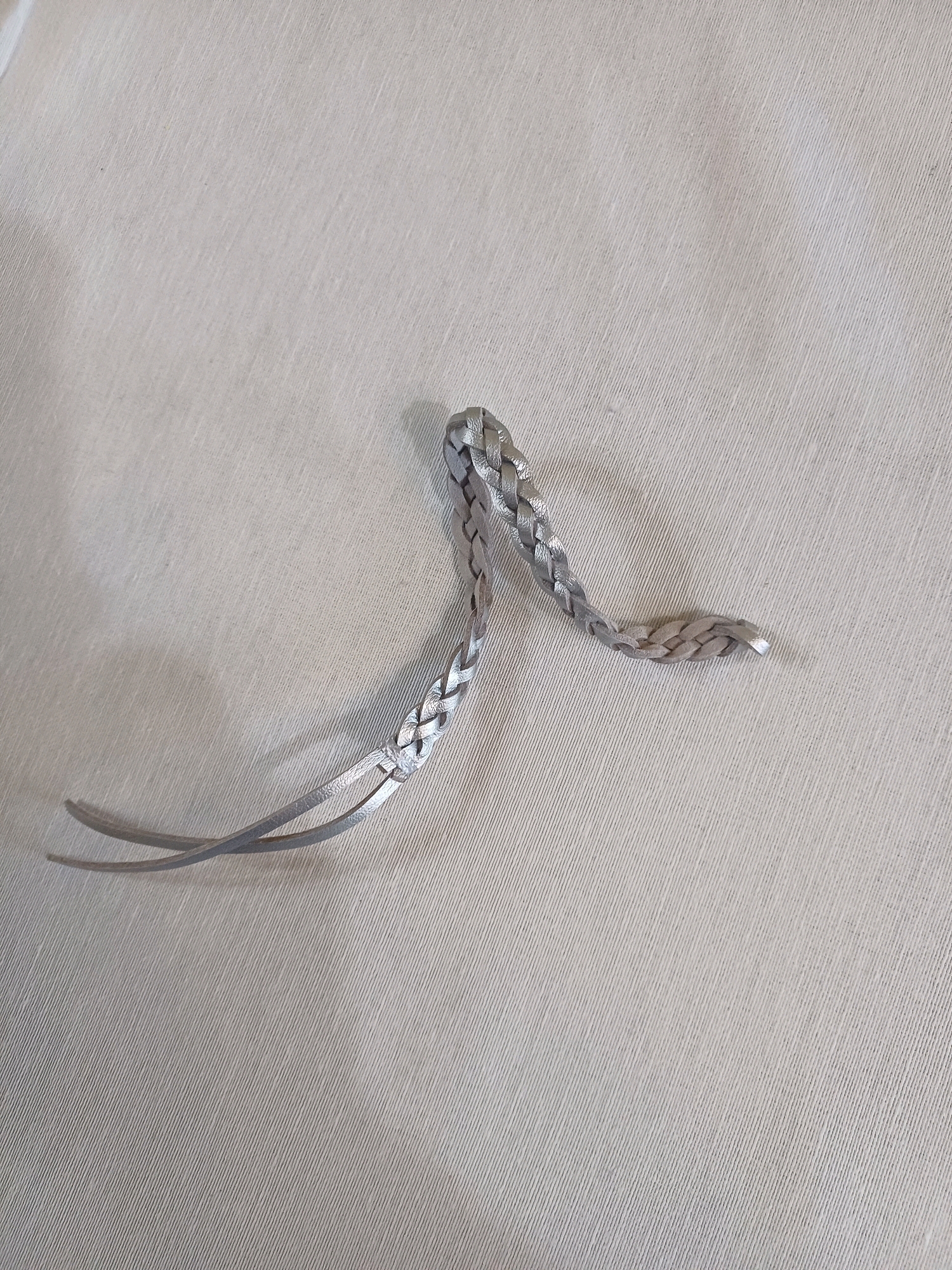A close-up, high-angle shot shows a braided silver-colored material, likely leather or faux leather, lying on a textured, light-colored fabric surface. The material is arranged in a curved shape, with one end appearing to split into two thinner strands that cross over each other. The braiding is tight and consistent, with a metallic sheen. The fabric background has a ribbed texture and a neutral tone, possibly a tablecloth or a piece of clothing. There are no people, and the scene appears to be an indoor still life, devoid of any context suggesting a location in Cheshnegirovo, Bulgaria, or any urban setting.