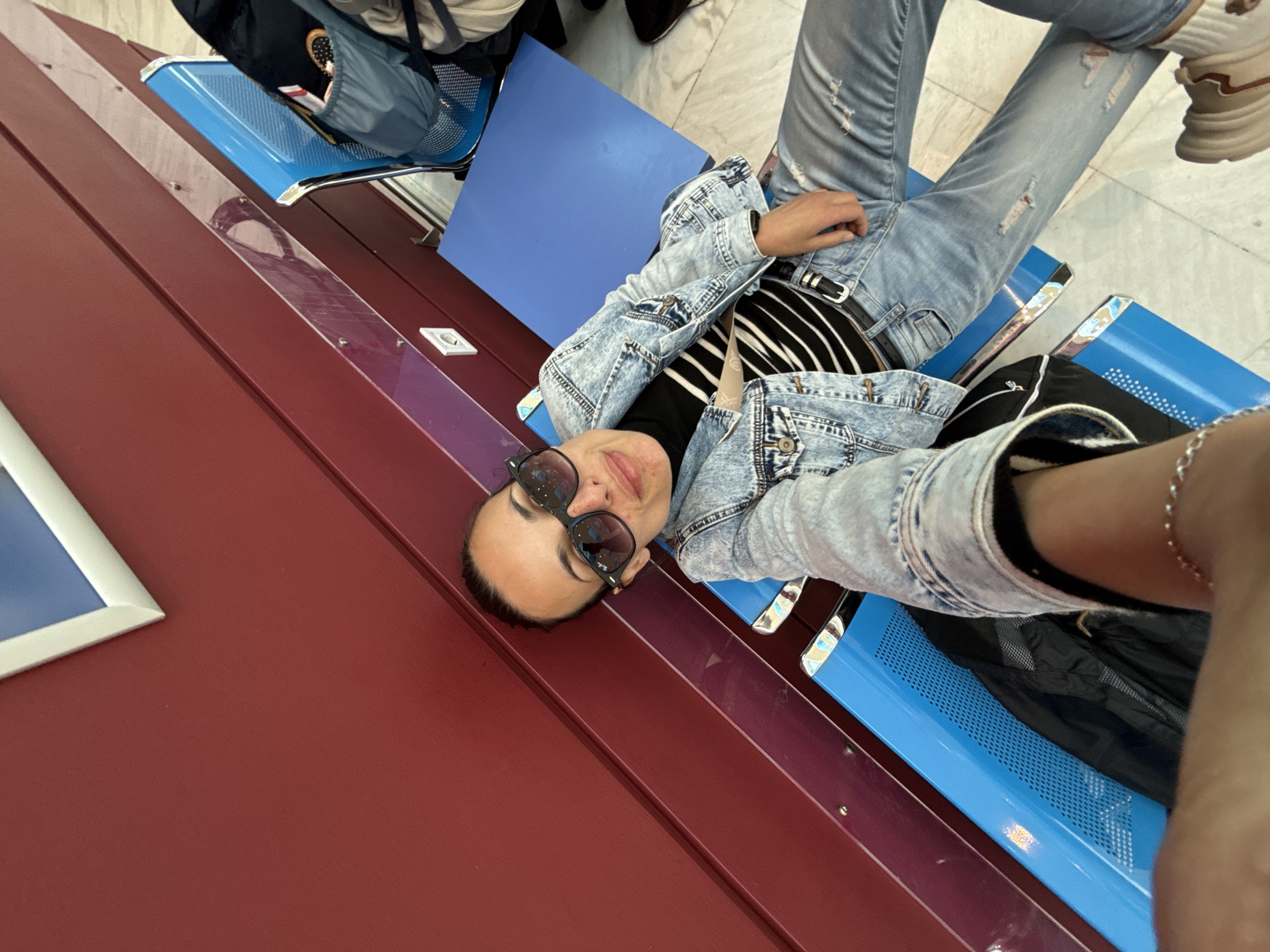 A person wearing a denim jacket and jeans is lying across blue airport-style seating in a building, possibly an airport or waiting area. The seating is red on one side. The person is wearing sunglasses and has a striped shirt underneath the jacket. The location cannot be verified as Mouzouras, Greece.