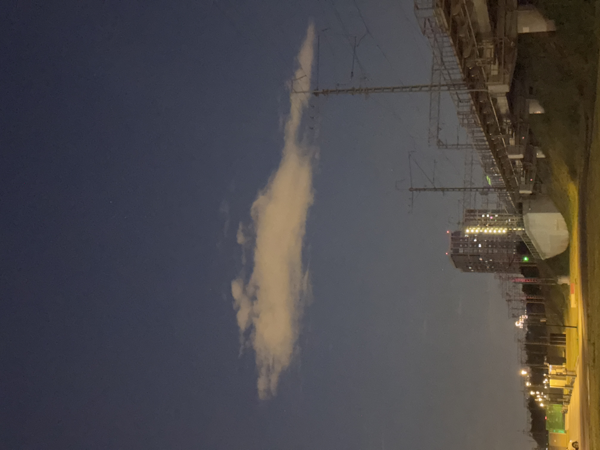 The image depicts a nighttime scene in a Moscow, Russia urban environment, likely on the outskirts. The sky is a deep, dusky blue with a single, elongated, illuminated cloud stretching horizontally across the upper third of the frame. Below the cloud, a tall, modern building with many illuminated windows stands prominently, its lights casting a warm glow. To the right of the building, a complex of railway infrastructure is visible, including an elevated railway bridge with multiple tracks, overhead electrical lines supported by tall metal poles, and supporting concrete structures. The bridge appears to be a significant piece of infrastructure, suggesting an active transportation hub.
In the foreground and mid-ground, streetlights illuminate a paved area, likely a road or parking lot, with yellow light. A few sparse street lamps are also visible along the road. A patch of grassy embankment is present to the right of the paved area, leading up to the railway bridge. The overall atmosphere is one of a developing or industrial area within the city, with a blend of residential and transportation infrastructure. There are no people visible in the image. The lighting suggests it is either twilight or night, with artificial lights from buildings and streetlamps dominating the scene. The cloud's illumination implies it is catching light from sources below, possibly city lights or reflected moonlight.