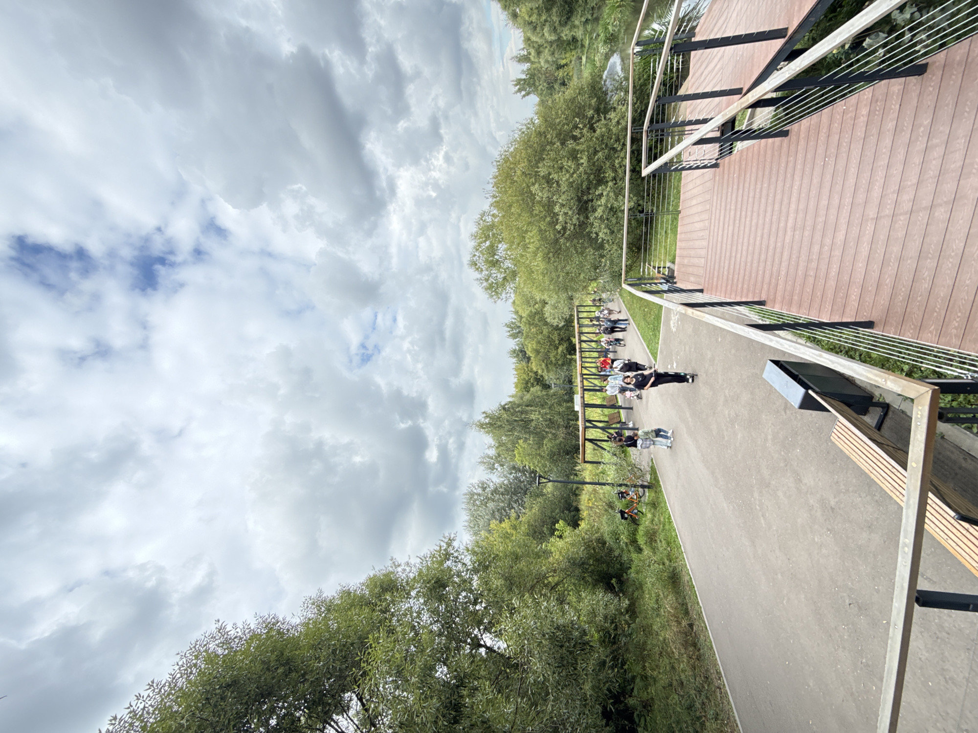 The image depicts a park scene with a paved walkway and a wooden observation deck. In the background, there is a line of trees and a body of water, suggesting a natural or semi-natural environment within a city. Several people are visible on the walkway, some walking and others on bicycles or scooters, indicating recreational activity. There is also a covered structure with seating, likely a pavilion or rest area. The sky is cloudy, suggesting overcast weather.
The provided information indicates the location is in Moscow, Russia. Based on the visual cues of a well-maintained park with recreational facilities, it is likely a public park or nature reserve within the city, designed for public use and leisure activities. Specific details about the exact park name or district are not evident from the image alone.