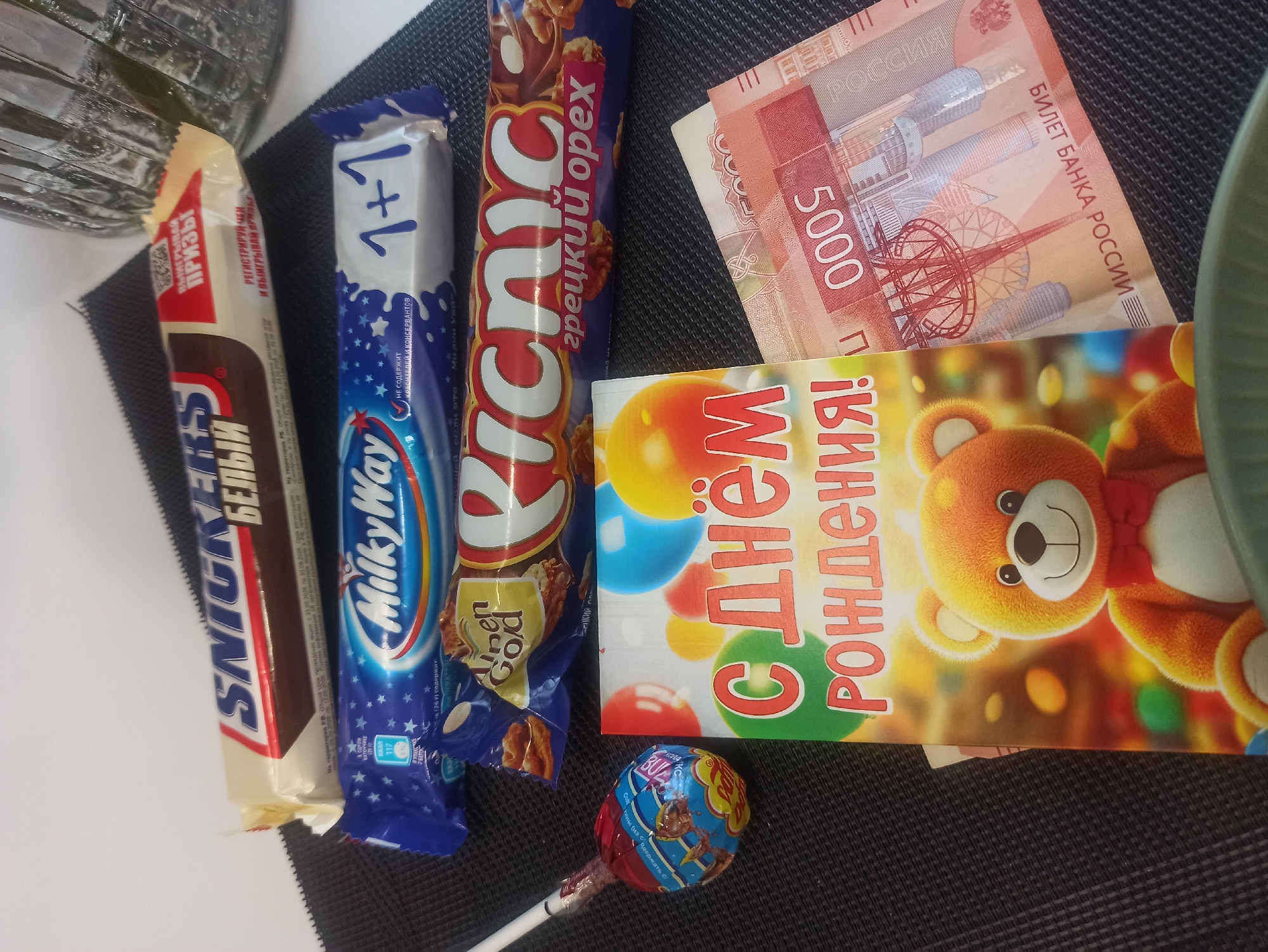 The image shows a flat surface covered with a textured, dark-colored material. On the surface are several objects including a Snickers White chocolate bar, a Milky Way 1+1 chocolate bar, a Picnic chocolate bar with "грецкий орех" (walnut) written on it, a lollipop, a birthday card with a bear on it, and a stack of Russian 5000 ruble banknotes. The birthday card reads "С днём рождения!" (Happy Birthday!). The location in the city of Kondratovo, Russia, is not determinable from the image. There are no people or actions visible in the image.