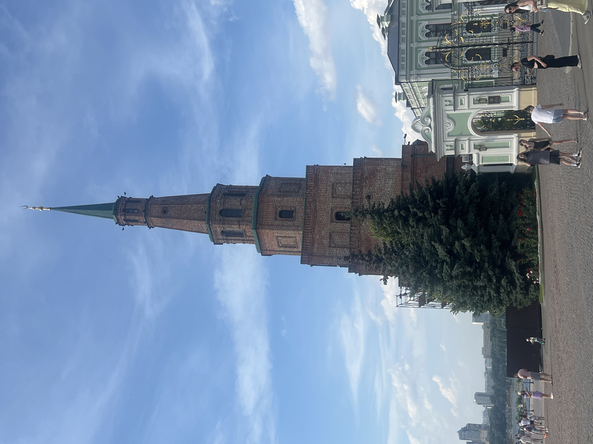 The image shows the Leaning Tower of Söyembikä in Kazan Kremlin, Kazan, Russia. The tower is a brick structure with a green spire topped by a crescent moon. The tower is partially obscured by a large evergreen tree in the foreground. To the right of the tower, there is an ornate green and white gate and a building with a green roof. Several people are visible in the foreground, including adults and children, some of whom are walking and others standing. In the background, there is a cityscape with several buildings and a body of water. The sky is blue with some scattered clouds. The ground in the foreground is paved with cobblestones.