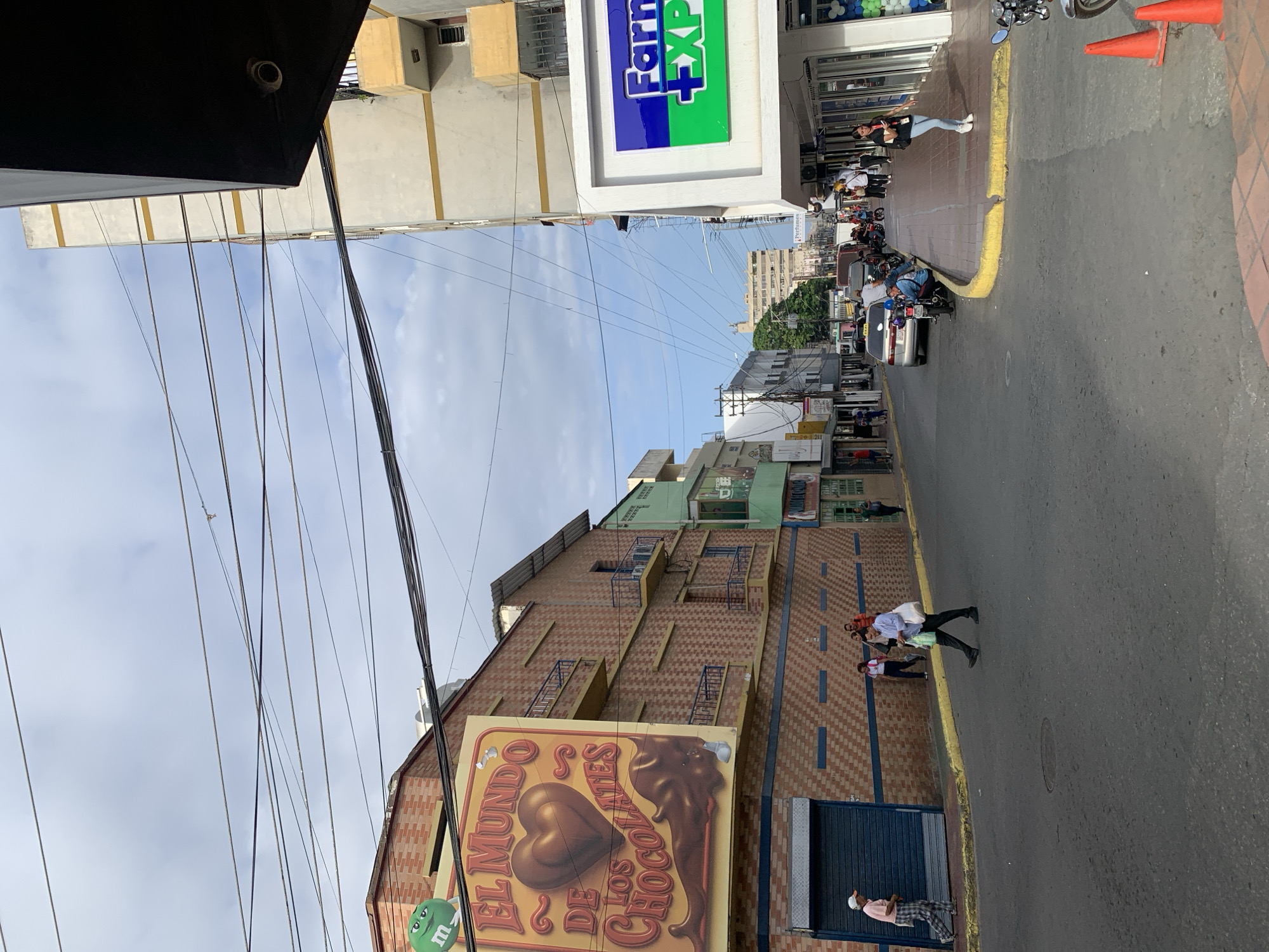This media file captures a vibrant street scene in Porlamar, Venezuela, under a bright, clear sky with scattered clouds. The image is oriented vertically, showing a mix of commercial and residential buildings lining a paved street.
On the left side of the street, a prominent building features a large, colorful sign for "EL MUNDO DE LOS CHOCOLATES" (The World of Chocolates), depicting a chocolate heart and melted chocolate. Below the sign, a green M&M character is visible. The building's facade above the sign is adorned with a brown and beige brick pattern, and multiple window-like structures with blue grates are spaced across it. Further down this side of the street, other buildings with varying architectural styles and colors, including light green and beige, can be seen.
On the right side, another multi-story building displays a large blue and green sign with "Farm" and "+XP" written on it, suggesting a pharmacy or similar retail establishment. People are seen standing and walking on the sidewalk in front of this building, some near parked motorcycles. Orange traffic cones are placed near the curb.
The street itself is asphalt with a distinct yellow line separating the roadway from the sidewalk. Several motorcycles are parked along the curb, and a silver car is also visible. People are engaged in daily activities; some are walking on the sidewalks, including one person in a light blue shirt carrying a white bag, and another in a pink shirt and plaid pants wearing a cap, standing near a closed blue corrugated door.
Numerous power lines and cables crisscross high above the street, a common sight in urban areas. The overall impression is that of a bustling daytime street in a city, likely in the late morning or early afternoon given the bright, even lighting.