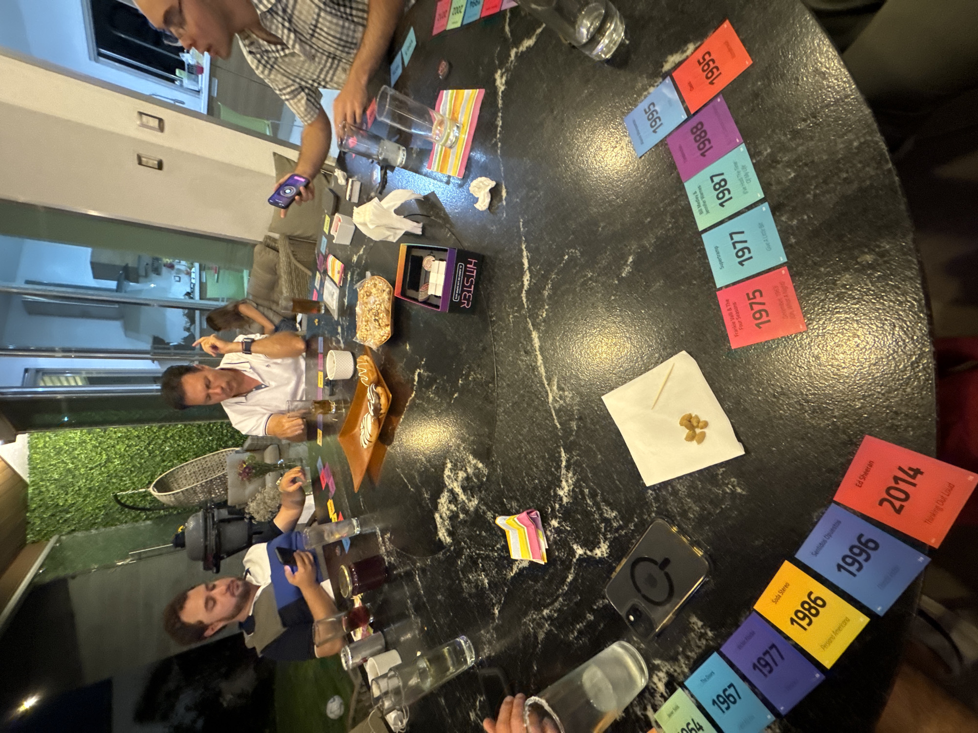 A group of people are gathered around a round, dark-colored marble-patterned table playing a board game, likely the "Hister" game, as evidenced by the game box on the table. The game involves placing cards with different years on them in chronological order. The cards visible have years ranging from 1964 to 2014. The table also has glassware, a napkin with food items, and some decorative items. Three men are clearly visible, two in shirts and one in white. One is operating a phone and sitting on the floor, leaning against the table. The setting appears to be indoors, potentially in a well-lit residential space that could be a house or an apartment with large windows facing a patio with greenery. Without additional information, there is no basis to associate this scene with San Bernardino Tlaxcalancingo, Mexico.