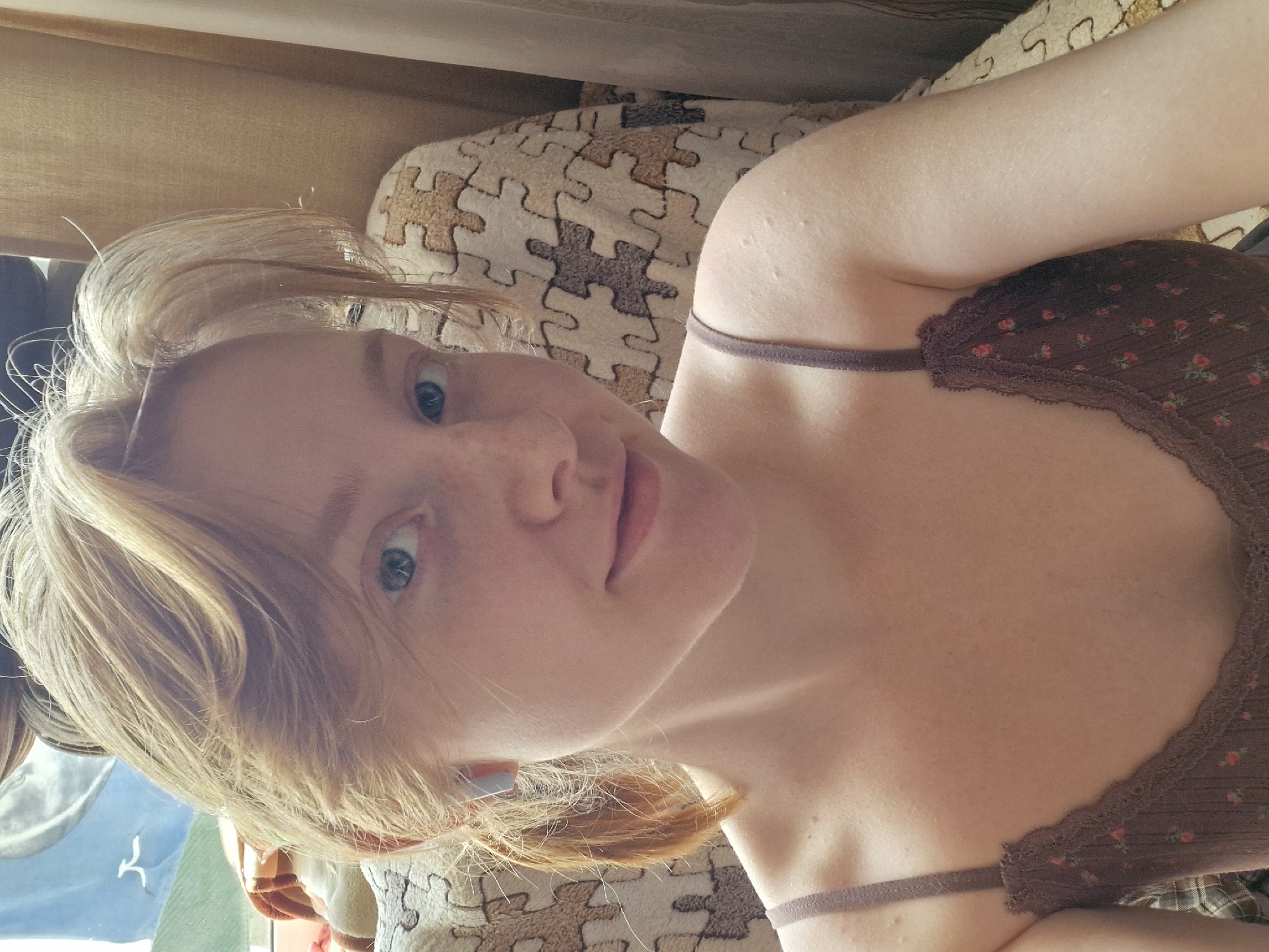 A young woman with fair skin and light blonde hair is taking a selfie. She is wearing a brown floral print tank top. The background includes a beige and brown puzzle-patterned fabric, possibly a blanket or pillow, and what appears to be a tan-colored curtain or shade. The location in Yekaterinburg, Russia is not determinable from the image.
