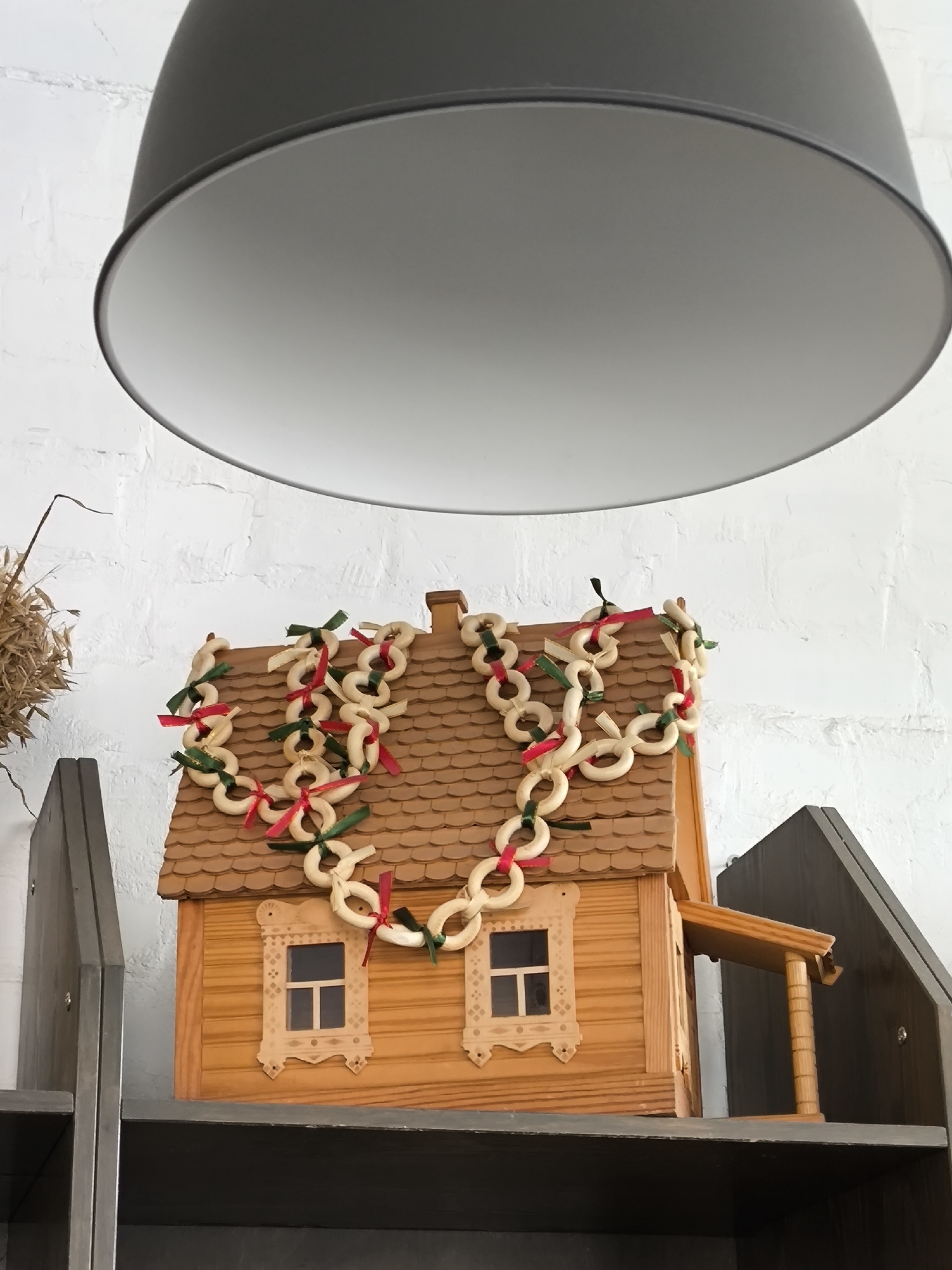 A miniature wooden house with a tiled roof is displayed on a shelf against a white brick wall. The roof is decorated with a garland of ring-shaped bread products, possibly bagels or pretzels, interspersed with red and green ribbons, forming a heart shape. The house has two windows with decorative carved wooden frames. A dark gray lampshade hangs over the top of the image, casting a shadow. To the left of the house, a bundle of dried grass is visible. The scene appears to be indoors, possibly in a home or a shop in the city of Tver, Russia, given the stylistic elements of the house decoration.