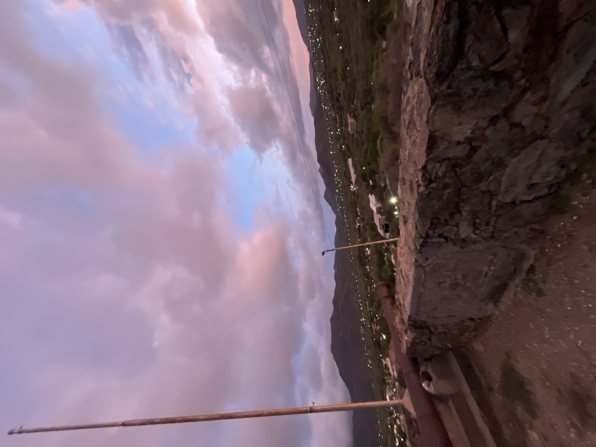 The image depicts a twilight view of Santa Ana, Venezuela, from an elevated vantage point, likely a historical site. In the foreground, a weathered stone wall forms a barrier, with a large, dark, cylindrical object, possibly an old cannon barrel, resting on it and extending towards the left. To the left of the wall, a tall, wooden pole stands vertically. Beyond the wall, the landscape rolls out, featuring a sprawling town illuminated by numerous lights, nestled against the backdrop of dark, mountainous terrain. The sky is awash in the soft hues of sunset, with clouds painted in shades of pink, purple, and pale blue, suggesting a calm evening atmosphere.