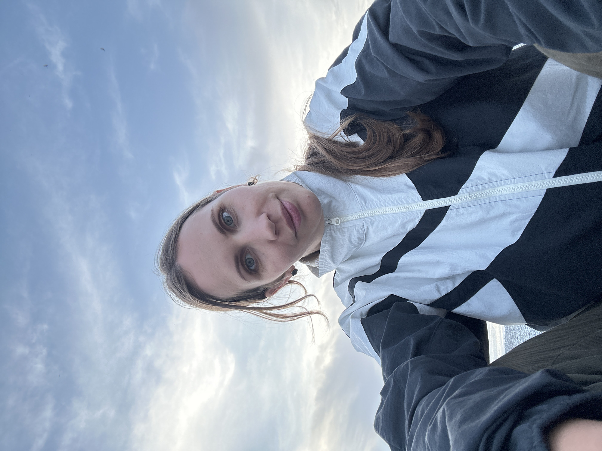 A low-angle selfie shows a Caucasian woman with light brown hair and blue eyes, wearing a black and white jacket, against a backdrop of a cloudy sky. The setting appears to be outdoors, given the sky in the background. Without additional visual information or metadata, the specific location in Fethiye, Turkey, cannot be determined.