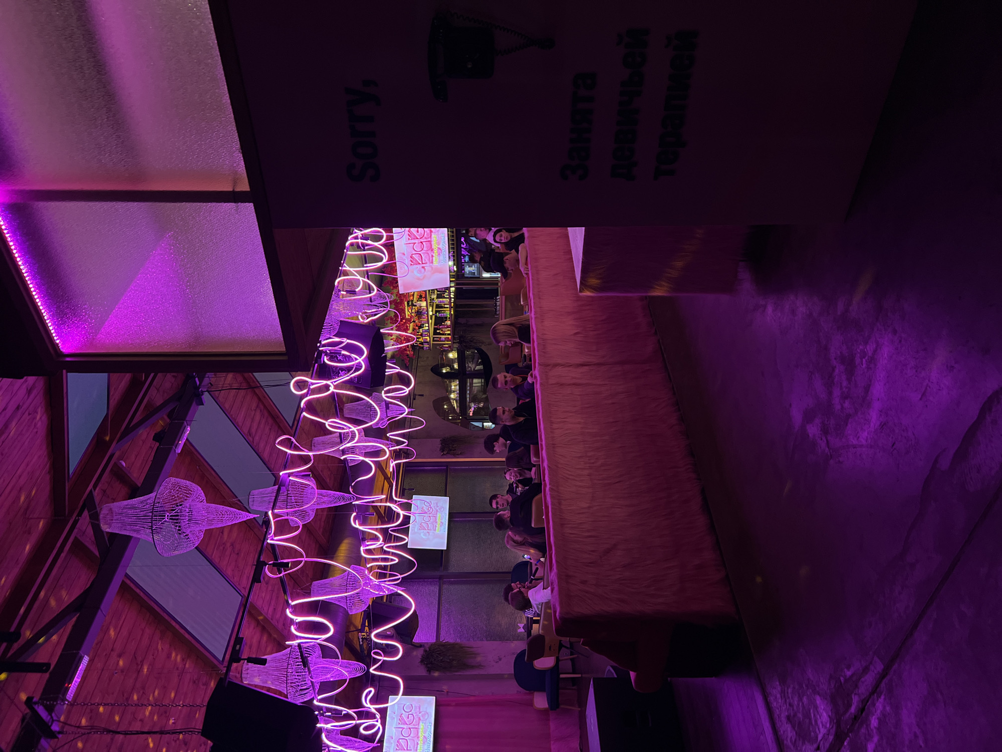 This image captures the vibrant interior of an establishment, likely a bar or club, bathed in striking purple and magenta neon lighting. The scene is set in the evening or night, indicated by the sole reliance on artificial light sources.
The main subjects are several people seated along a long, low table or bar that features a distinctive fluffy, light brown or pinkish-brown texture. They appear to be socializing, with some faces visible and engaged in conversation.
Prominently featured are decorative elements including a long, winding neon light fixture casting a bright magenta glow across the ceiling. Below these neon lights, several elegant, possibly metal-wire or beaded chandeliers hang. The ceiling itself is a mix of exposed wooden beams and glass panels, with LED strip lighting along the edges of some panels.
On the upper right side of the frame, a dark sign with white text is visible. It reads "Sorry," followed by a depiction of a vintage black telephone. Below the telephone, the Russian text "Занята девичьей терапией" is displayed, which translates to "Busy with girl therapy." Further back in the venue, two small screens are visible, one clearly showing the numbers "1994." A large dark speaker stands in the bottom right corner.
The overall atmosphere suggests a trendy and lively social gathering spot, characterized by its unique lighting, decorative elements, and modern architectural features.