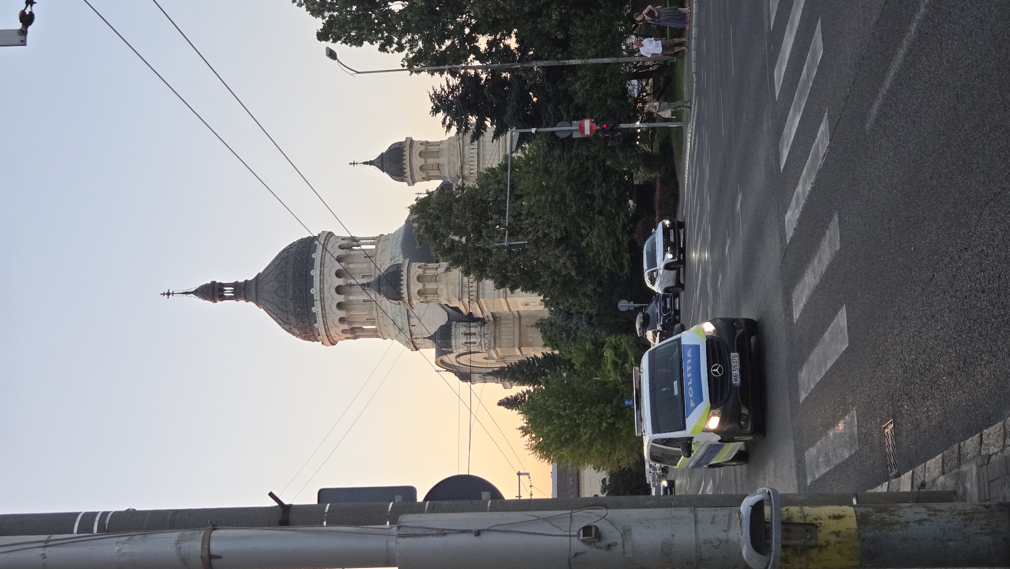 The image depicts a street scene in Cluj-Napoca, Romania, dominated by the Transfiguration Cathedral. In the foreground, a white police van with "POLITIA" written on it is driving across a pedestrian crossing. The van has its headlights on, and a person is visible in the driver's seat. Behind the police van, other vehicles are driving on the road, including a black SUV and a white car. In the background, the large Transfiguration Cathedral with its distinctive dome and multiple smaller domes is visible. Lush green trees surround the cathedral and line the street. The sky is a soft, pale orange, suggesting either dawn or dusk. Several people are standing on the sidewalk to the right of the image, observing the scene. The overall environment appears to be a public street in an urban setting, likely during evening or early morning hours.