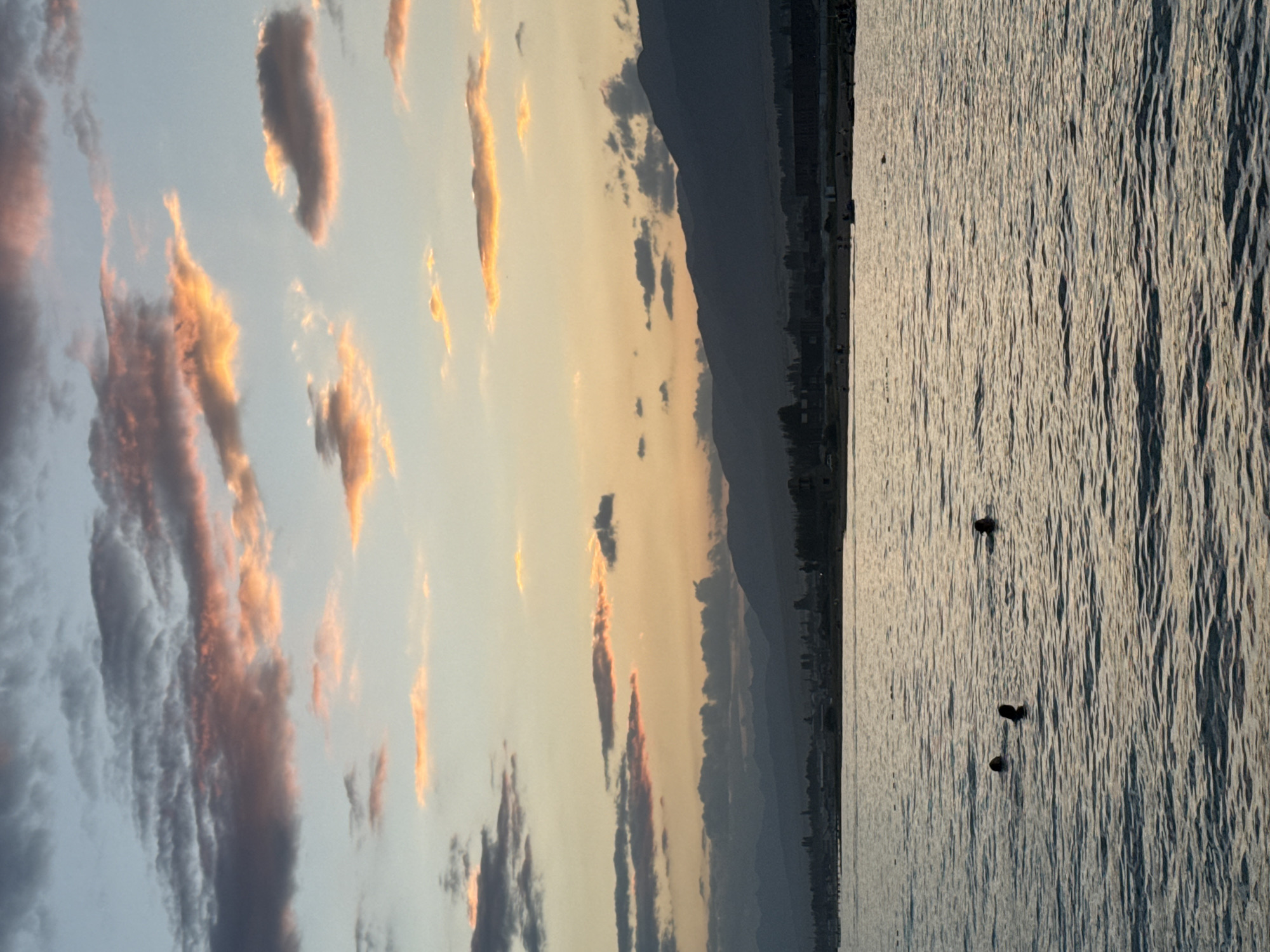 The image shows a body of water with three people swimming. The water is a calm, choppy surface reflecting the light. In the background, there is a distant shoreline with buildings and silhouetted mountains. Above the shoreline, the sky is filled with clouds tinged with orange and pink from a sunset. This scene is likely located in Sary-Oy, Kyrgyzstan, a resort town on the northern shore of Issyk-Kul Lake, known for its beaches and mountain views.