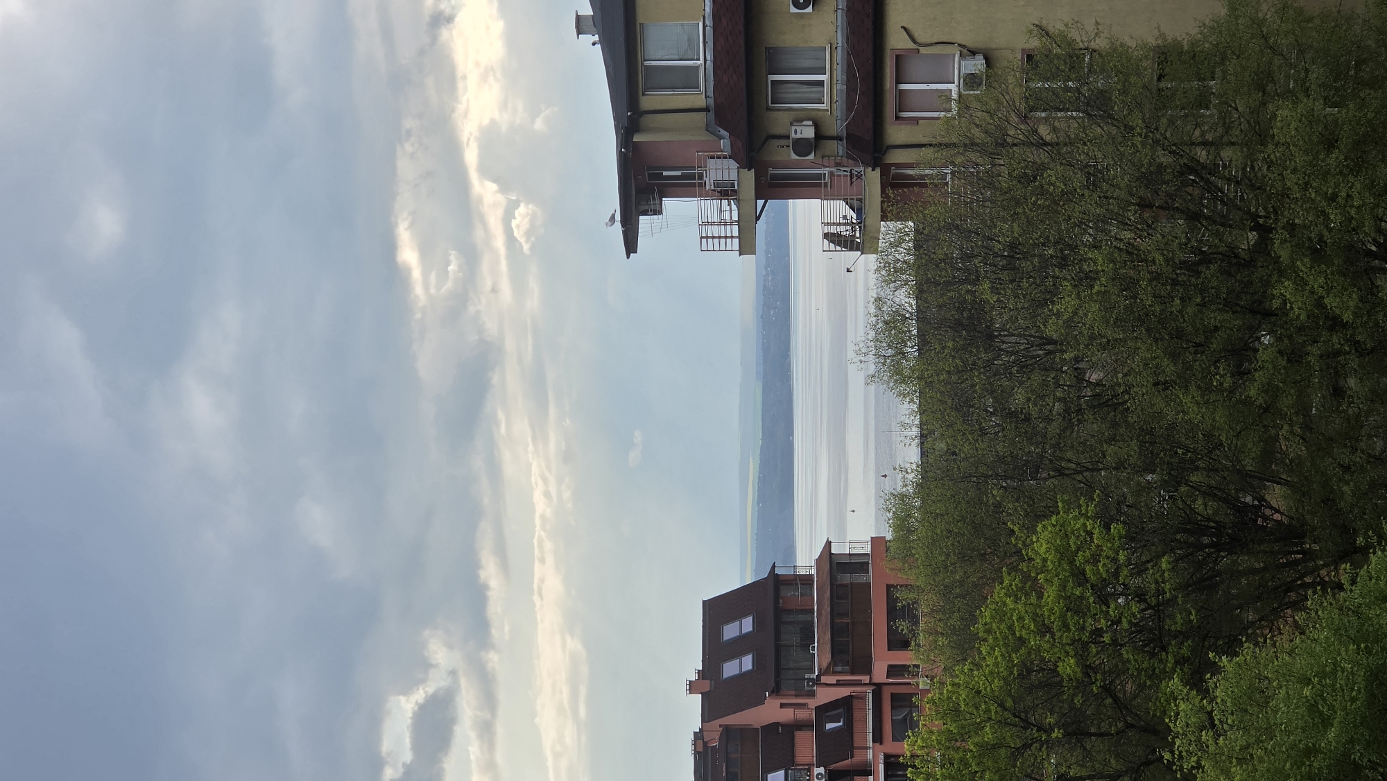 The image depicts a view of Varna Lake from a slightly elevated position in Varna, Bulgaria. On the left and right sides of the frame are buildings, one in a light yellow and red palette, and the other in shades of red. Mature trees with spring foliage are visible between and around the buildings. A body of water, Varna Lake, stretches across the center, with the opposite shore visible in the distance. The sky above the lake is filled with light, wispy clouds.